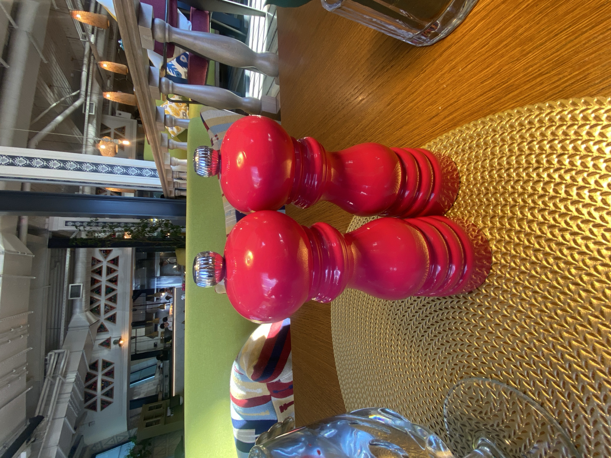 Two red ceramic salt and pepper grinders with chrome tops sit on a gold woven placemat on a wooden table. The grinders are positioned side-by-side in the foreground. To the left of the grinders, a portion of a clear glass is visible. In the background, a restaurant interior is apparent with green upholstered seating, colorful striped pillows, and decorative triangular patterns on a white wall. The restaurant appears to be in the city of Solnechnoye, Russia.