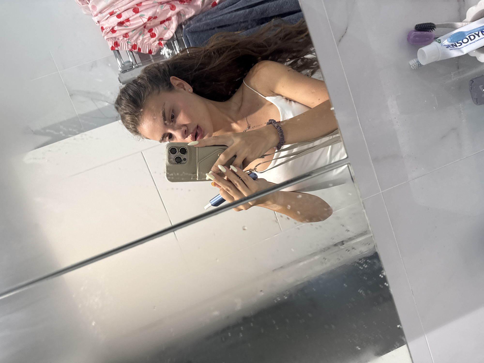 A young woman with long, dark, wavy hair is taking a mirror selfie in a modern bathroom. She is wearing a white spaghetti strap tank top and has long, light-colored fingernails. She is holding a smartphone with a light-colored case in her right hand, and in her left hand, she is holding a blue electric toothbrush. She is looking at her reflection in the mirror, with her lips slightly parted.
The bathroom features white marble-patterned tiles on the walls and a white countertop. A towel rack with a pink robe adorned with cherries and a dark blue towel is visible in the background. On the countertop, a tube of Sensodyne toothpaste and two toothbrushes, one purple and one black with grey bristles, are present. The mirror has water spots on its surface. The image is taken from a slightly low angle, looking up at the woman. The location in the city of Throop, United Kingdom, is not ascertainable from the image provided.