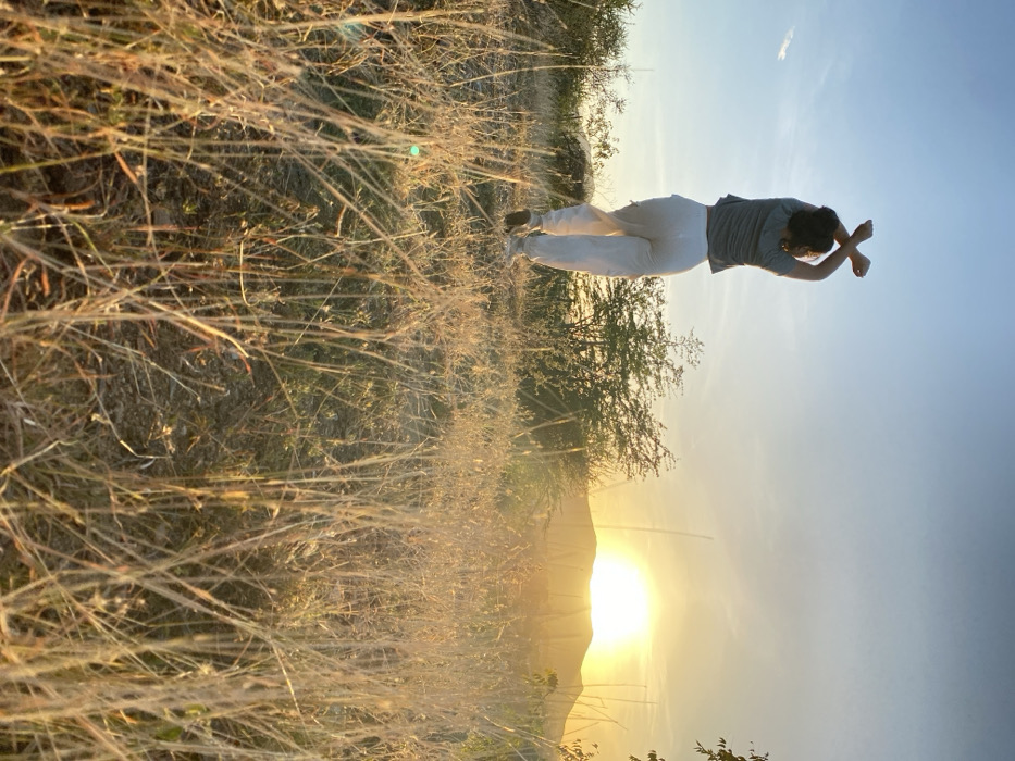A woman wearing jeans and a gray t-shirt poses with her arms crossed at her wrists against a landscape of tall, dry grass. The sun is low on the horizon, creating a warm, golden glow. Trees and a hill can be seen in the background against a clear sky. The image is taken from a low angle, emphasizing the height of the grass and the vastness of the sky.