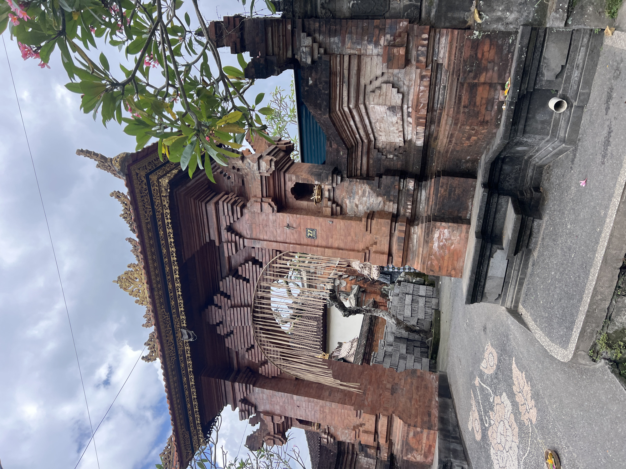 This image depicts an ornate, traditional Balinese temple entrance in Ubud District, Indonesia. The structure is constructed primarily of weathered red brick with intricate golden carvings adorning the roofline and archways. A canopy of bamboo hangs across the entrance, and a small, bare tree stands centrally within the archway. Piles of grey concrete blocks are visible behind the tree, suggesting ongoing construction or renovation. The ground leading up to the entrance is paved with asphalt and features a decorative mosaic pattern. A small offering basket, typically used in Balinese religious practices, rests on the left side of the pathway. Lush green foliage, including pink flowers, frames the upper right portion of the structure. The sky above is overcast with dramatic clouds.