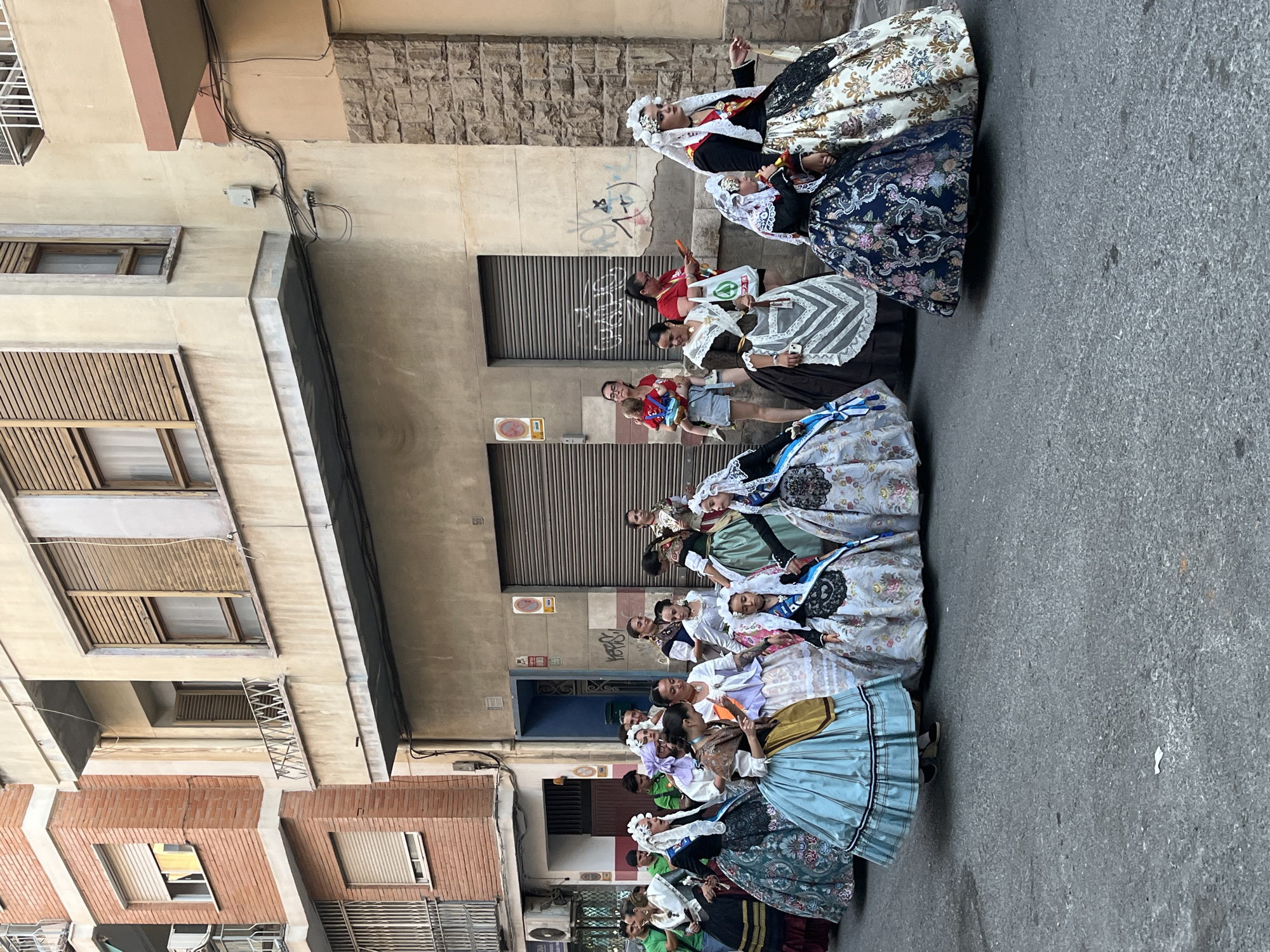 A group of women in traditional Valencian dress, consisting of voluminous skirts, blouses, and elaborate headdresses, are walking down a street. The women appear to be participating in a procession or parade. In the background, there are buildings with cream-colored facades, some with exposed brickwork and shutters. There are also some windows and balconies visible. A few other people, not in traditional dress, are also present on the periphery of the group. The street is paved with asphalt and appears to be a typical urban setting. The lighting suggests it is daytime. This scene is consistent with events that take place in Alicante, Spain, where such traditional attire is worn during festivals and celebrations.