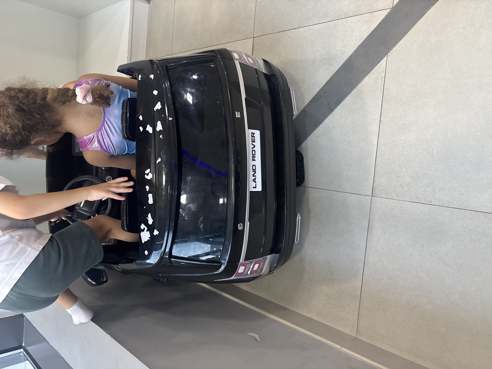 A high-angle shot captures two young children inside a black toy Range Rover. The child on the left, partially visible from the knees down, wears a white t-shirt and dark green shorts with white socks. This child is climbing into the toy car from the left side, with one leg extended upwards. The child on the right, facing away from the camera, has curly brown hair tied with a pink and white scrunchie and wears a shimmery pink, purple, and blue leotard. They are seated in the driver's seat, with their hands visible on the steering wheel and dashboard. The toy car is parked on a tiled floor with light grey and white tiles. The rear of the toy car features a "LAND ROVER" emblem. The scene is likely indoors, possibly in a retail or play area, given the clean tiled flooring and the presence of a wall with a lighter grey section and a dark grey strip. The image lacks any specific Dubai landmarks, thus its location within the city cannot be determined from the visual information provided.