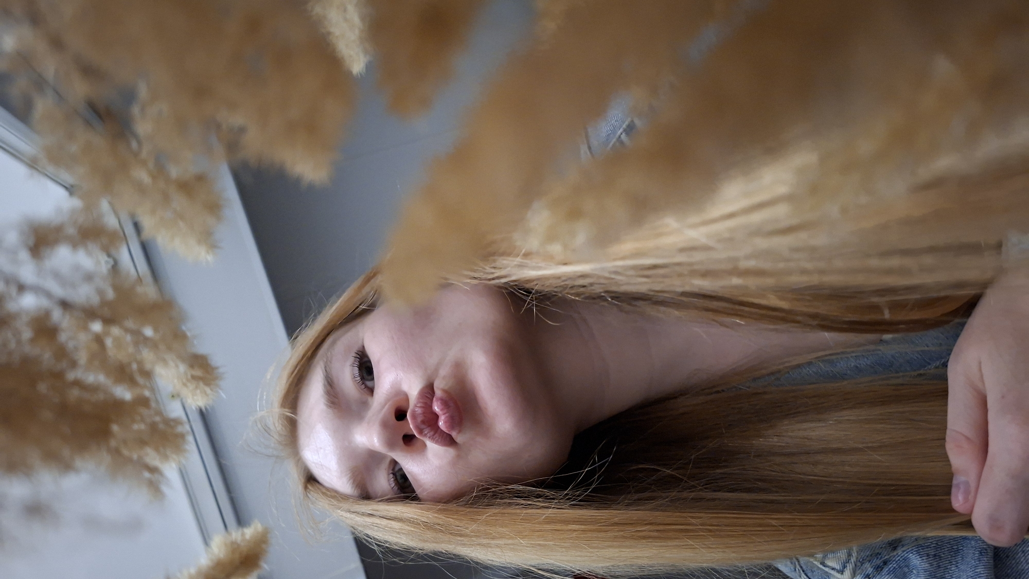 A young woman with long, blonde hair is holding her head down and puckering her lips towards the camera. She is wearing a denim jacket. Her left hand is visible in the lower left corner of the frame, resting on her chest. The background appears to be an indoor setting with a light-colored ceiling and some decorative dried grass or reeds hanging from above. There is no indication of the specific location in Chaikovsky, Russia, other than that it is likely an indoor space.
