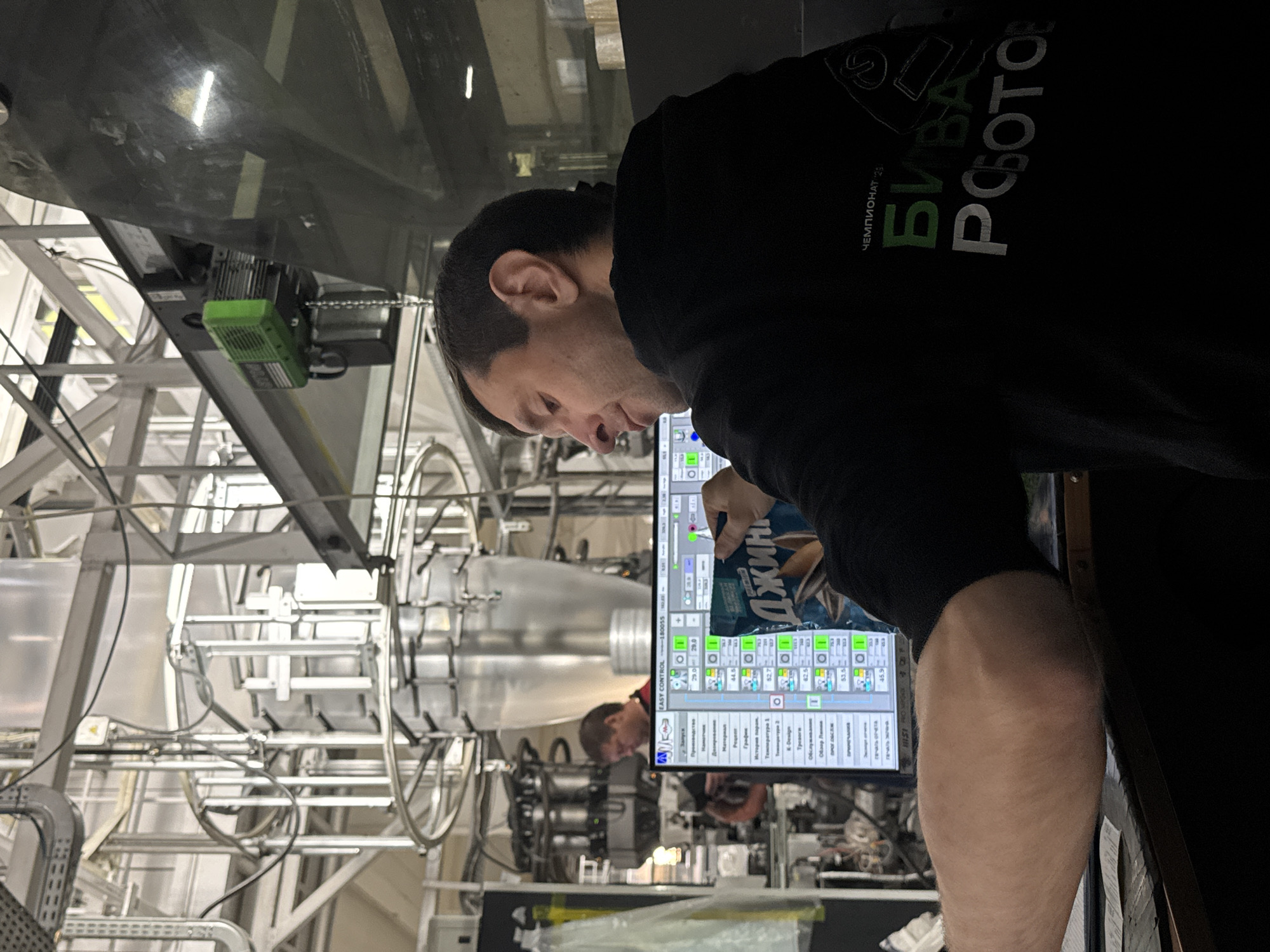 A man in a black sweatshirt with the text "ЧЕМПИОНАТ 2023 БИВА РОБОТОВ" is standing in front of a computer screen. The screen displays a control panel for a manufacturing process. In the background, there is industrial machinery, including pipes, tanks, and structural supports. The setting appears to be a factory floor, possibly located in Perm, Russia, based on the text on the sweatshirt. Another person is visible in the reflection on the screen. The man is holding a bag of "Джин" chips. The image is oriented sideways.