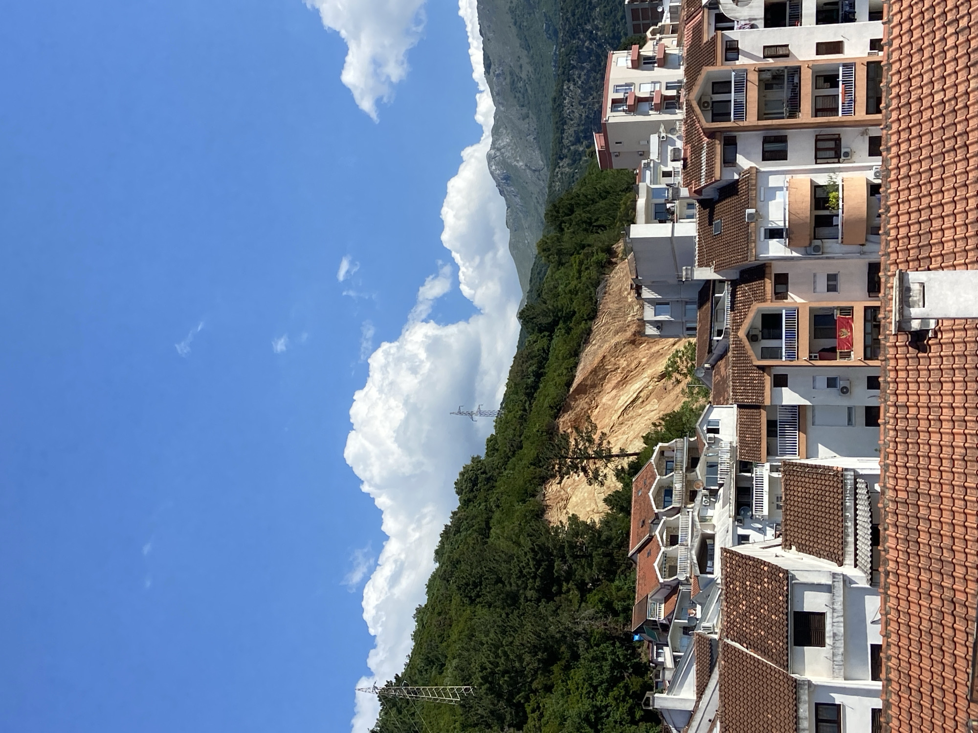 The image shows a view in Budva, Montenegro. Modern, white residential buildings with orange tile roofs are constructed along a hillside. Green trees and a tan dirt cliff are interspersed among the buildings. Tall, grey mountains rise in the background. The sky is blue with large, white cumulus clouds. A silver power transmission tower can be seen on the hillside.
