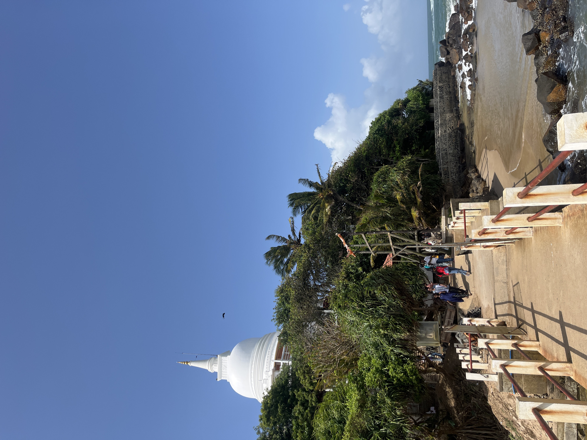 This is a wide-angle, low-angle outdoor photograph of the Japanese Peace Temple (also known as the Japanese Dagoba or the Japanese Peace Pagoda) located on a hill overlooking the ocean in Matara, Sri Lanka. The white, stupa-shaped temple with a golden spire dominates the upper left portion of the frame. It is surrounded by lush green trees and vegetation.

In the foreground, a concrete walkway with red and white railings leads towards the temple. Several people are visible walking on the path, some heading towards the temple and others in the opposite direction. They are casually dressed. To the right of the walkway, there is a small sandy cove with rocky formations and the ocean. Waves are breaking on the shore. The sky is a clear, vibrant blue with a few scattered white clouds. A small black bird is in flight in the upper center of the image. The overall impression is a sunny, tropical day at a serene coastal location.