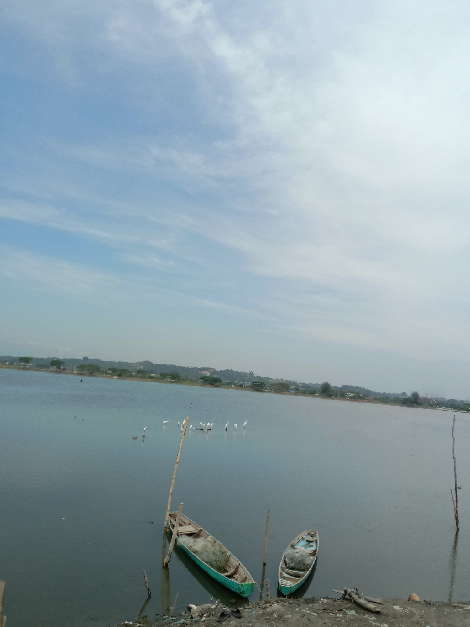 The image shows a body of water with two small boats near the shore. The boats appear to be wooden fishing boats. There are several white birds, likely herons or egrets, standing in the water in the middle distance. The far shore has vegetation and buildings, suggesting a populated area. The sky is overcast with scattered clouds. The location is Lhokseumawe, Indonesia, based on the image's metadata. The environment appears to be a coastal or estuarine area used for fishing. There is some debris on the shore. There are vertical poles sticking out of the water near the boats.