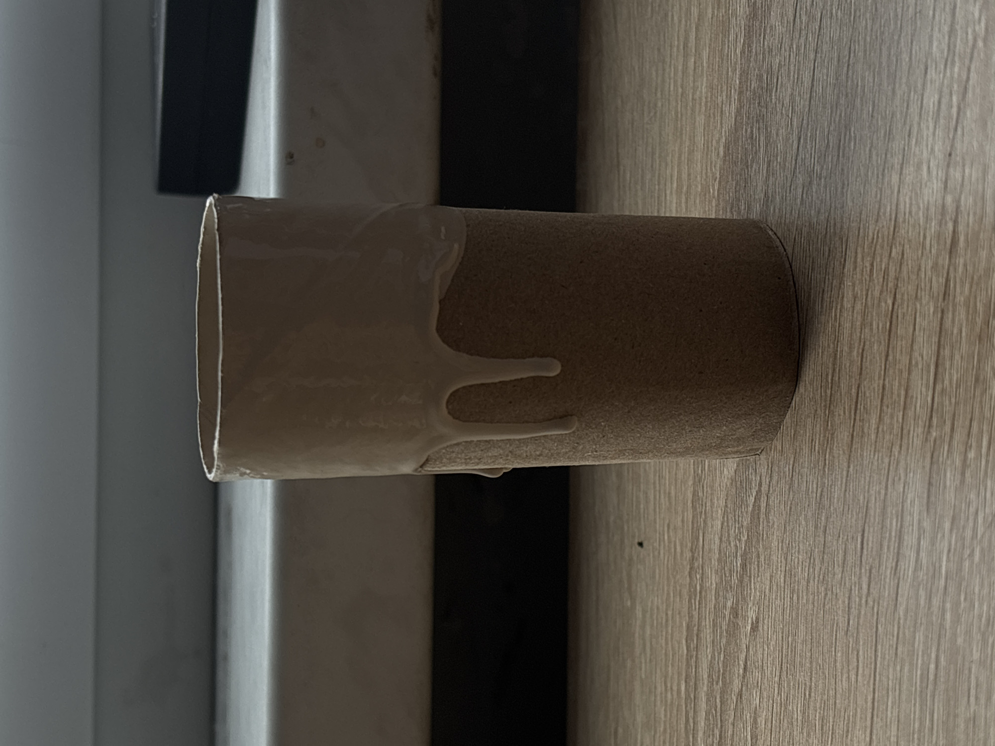 The image shows a close-up, eye-level view of a cardboard toilet paper tube on a wooden surface. The top edge of the tube has a beige liquid substance that is dripping down the sides in several places. The background is out of focus, with a lighter colored surface behind the tube and a dark, blurred area further back. There is also a dark rectangular object visible in the upper right background. The lighting appears to be coming from the left, casting soft shadows. The scene is indoors, on what appears to be a table or shelf. There is no indication of the location being in Saint Petersburg, Russia.
