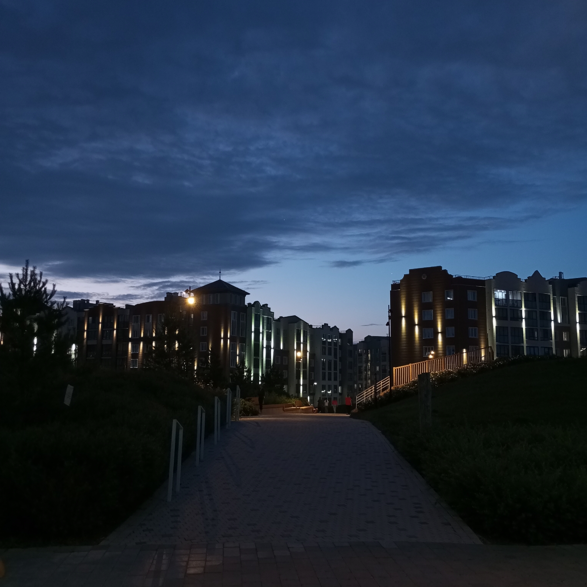 The image depicts a scene in Pervomaysky, Russia, at dusk or early evening. The sky is a deep blue, transitioning to lighter hues near the horizon, with some wispy clouds. The foreground is dominated by a paved walkway that leads towards a row of modern, multi-story apartment buildings. These buildings are illuminated by warm, vertical lights that highlight their facades, and some windows are also lit from within.
To the left and right of the walkway, there are grassy slopes covered in low-lying vegetation. Along the sides of the walkway, white posts are visible, likely part of a lighting system or railing. In the mid-ground, several people can be seen walking along the walkway or gathered near the buildings. Their figures are somewhat indistinct due to the low light conditions.
The overall atmosphere is calm and serene, suggesting a quiet evening in a residential area. The modern architecture of the buildings and the well-maintained walkway indicate a developed urban or suburban environment. The time of day is clearly indicated by the twilight sky and the artificial lighting of the buildings. There is no visible text in the image.
