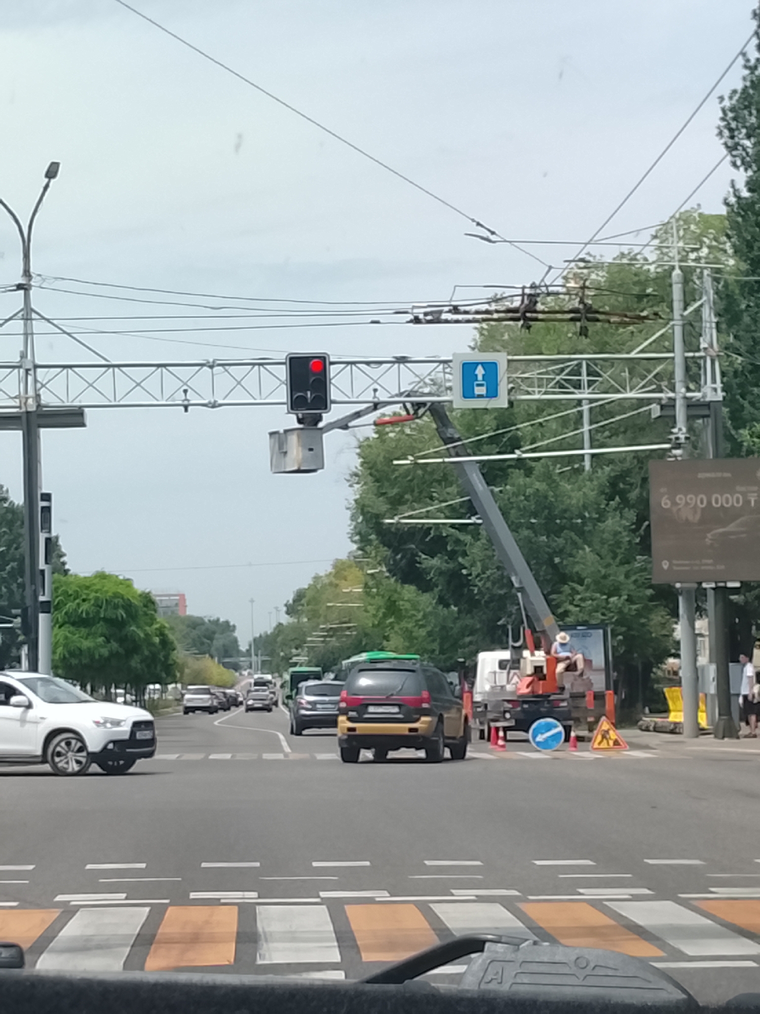 The image depicts a street scene in Almaty, Kazakhstan. A traffic light is visible, showing a red light. A sign with a blue background and a white arrow pointing upwards indicates a forward direction for vehicles. A construction crane is positioned over the road, with a person in a hat seated in the crane's basket, appearing to be working. Several vehicles are present, including a white SUV in the foreground on the left, and a yellow and black SUV further ahead. Road markings for a pedestrian crossing are visible. In the background, trees line the street, and buildings are partially visible. A large billboard with text in a foreign language and numbers is mounted on a pole. The sky is overcast.