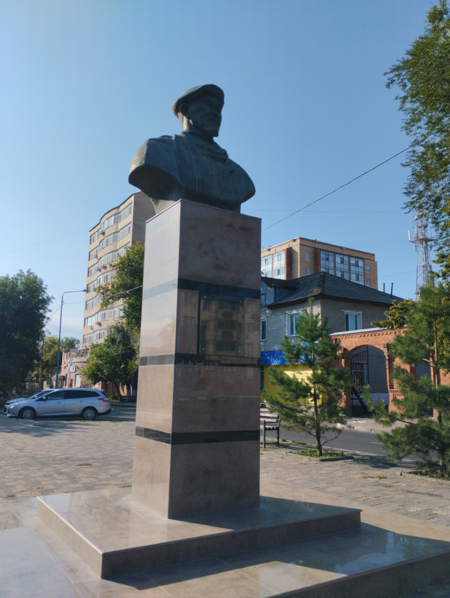 A bust monument of a man wearing a hat stands in a city square in Kostanay, Kazakhstan. The monument is made of dark bronze and is mounted on a tall, rectangular pedestal of polished granite. The pedestal has inlaid bands of black granite. Behind the monument, several buildings are visible. To the left is a modern, light-colored apartment building with many windows. To the right of that is a smaller, brick building with a gray roof, and further to the right, a modern building with a red brick facade. A telecommunications tower is visible in the background to the far right. The ground in front of the monument is paved with interlocking pavers. A silver station wagon is parked on the street in the middle ground to the left. Several small evergreen trees are planted in the square. The sky is clear and blue.