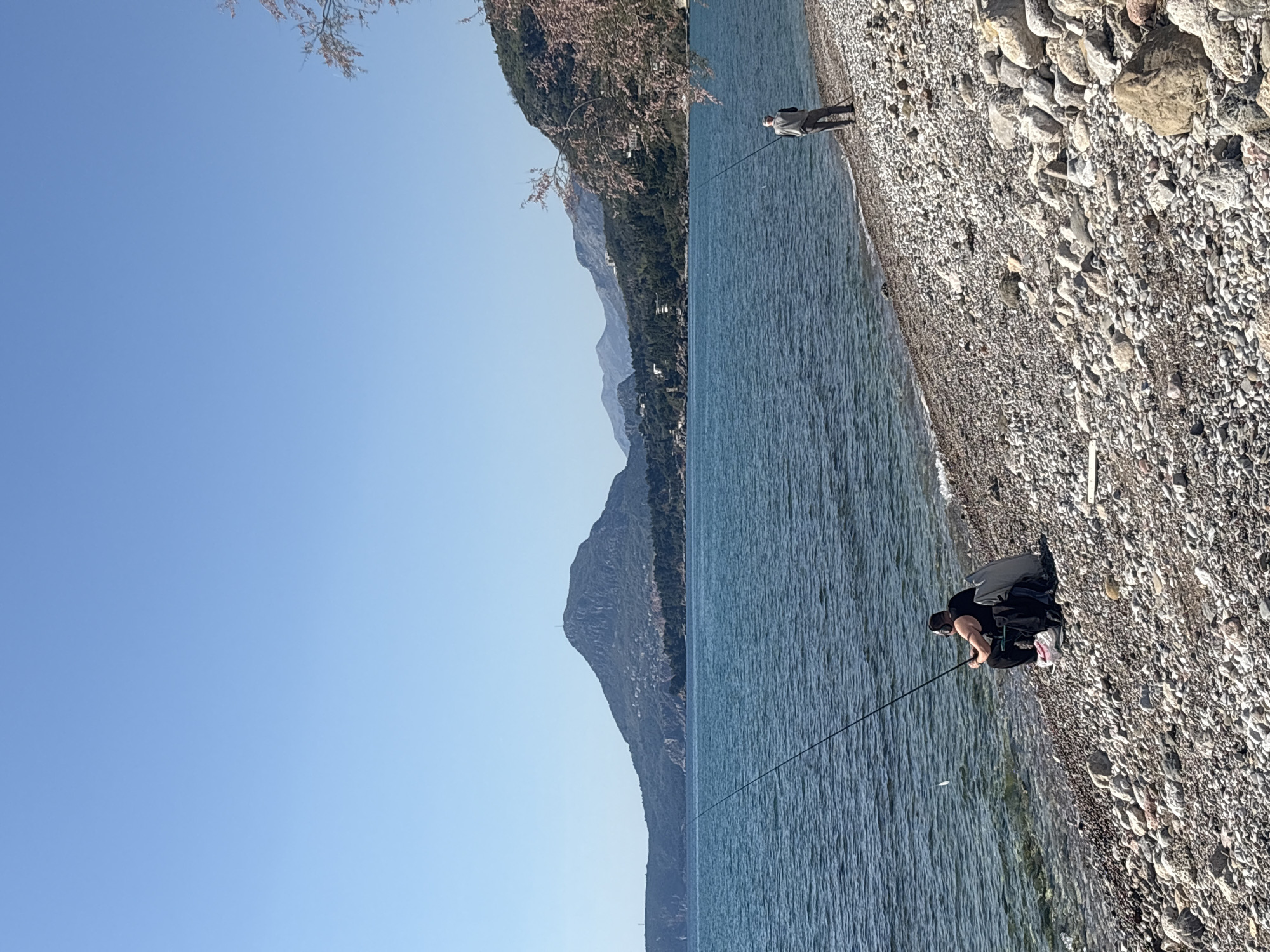 The image depicts a rocky beach scene at an unidentified location near Bar, Montenegro, showing two individuals fishing. The body of water appears to be a lake, bordered by mountains. One person is standing near the shoreline, holding a fishing rod. Another person is sitting or crouching on the rocky beach, also fishing. The terrain is covered with pebbles and rocks of varying sizes. The sky is clear and blue.