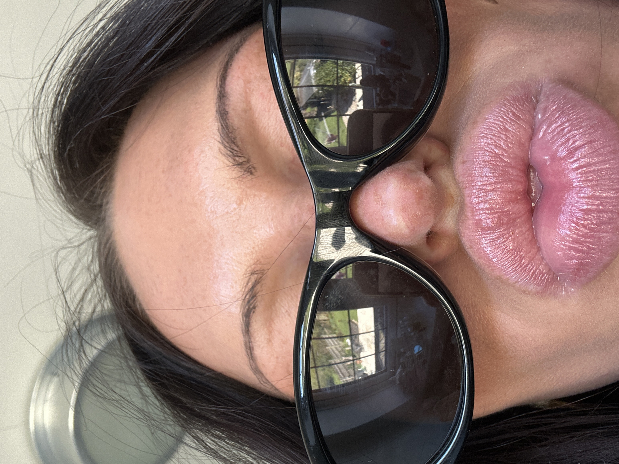 The image depicts a person's face wearing black sunglasses. The reflection in the lenses indicates an outdoor setting, possibly a garden or backyard. The person has dark hair and is wearing a glossy lip product. The nose is positioned between the lenses of the sunglasses. The background is blurred. It is not possible to determine the location in the city of Wealden, United Kingdom, from this image.