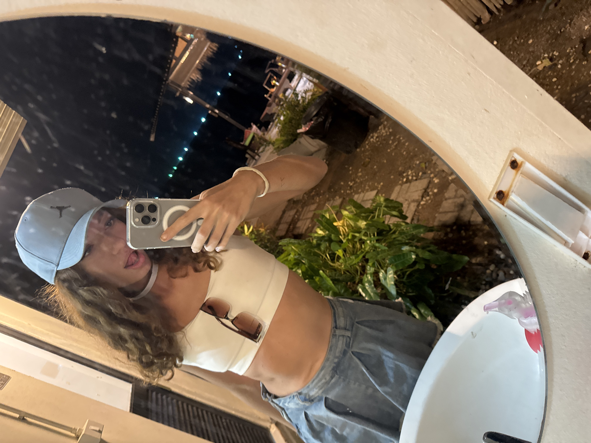 A woman stands in front of a mirror, taking a selfie with her smartphone. She is wearing a light-colored baseball cap, a white crop top, and blue jeans. Sunglasses are clipped to her top. The mirror reflects a nighttime scene with lights in the background, suggesting an urban setting, along with greenery and a sink in the foreground. The location is identified as Baan Bang Rak, Thailand.