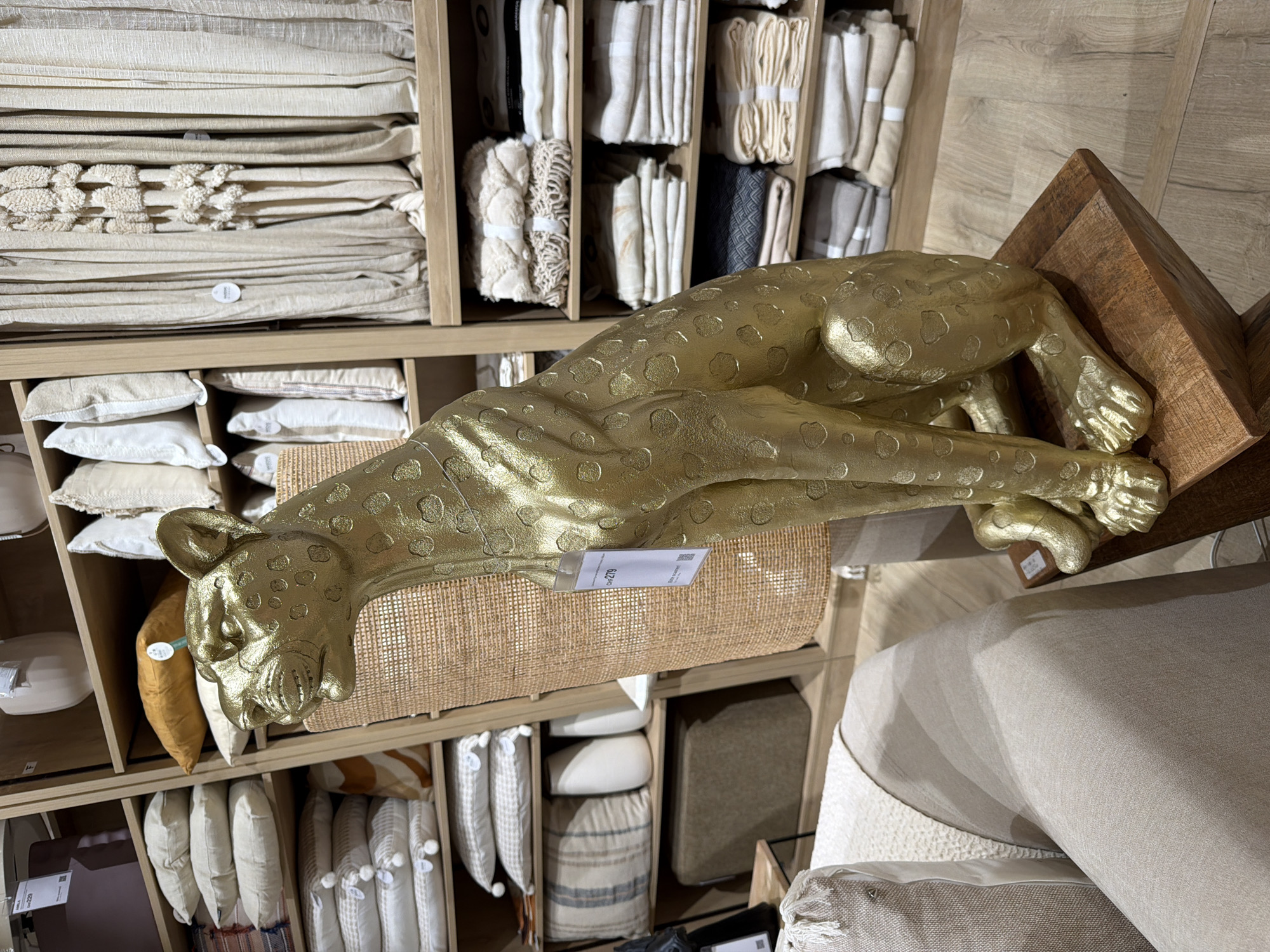 The image shows a golden leopard statue in what appears to be a retail store. The statue is in a crouched pose, and its surface is textured with circular spots. It is placed on a wooden block. Behind the statue, shelves are stocked with various home decor items, including folded textiles like blankets and pillows, as well as decorative vases. Some shelves are also lined with draped fabrics, possibly curtains. The overall environment suggests a well-organized display of home furnishings. The lighting is neutral, indicating an indoor setting. There are no people visible in the image. The location is described as Unknown, Unknown.