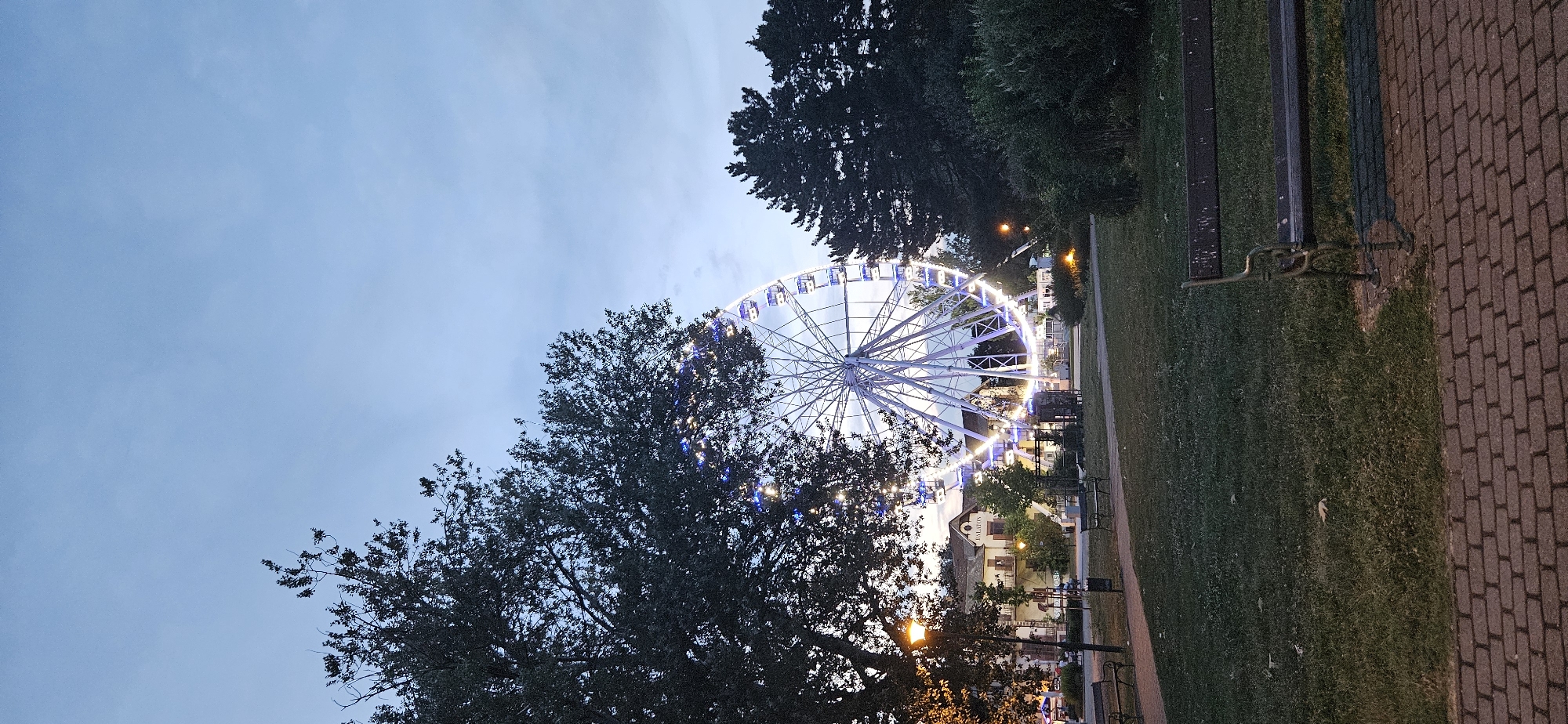 The image depicts a Ferris wheel illuminated with white and blue lights, set against a dusky sky. The Ferris wheel is situated in a park-like setting with grass, trees, and walking paths. In the background, a building with the word "BALATON" visible on its facade is present. Several park benches are visible in the foreground and midground. The scene appears to be in the evening or at dusk, judging by the lighting conditions and the illuminated Ferris wheel. This location is identified as Keszthely, Hungary, specifically near Lake Balaton.