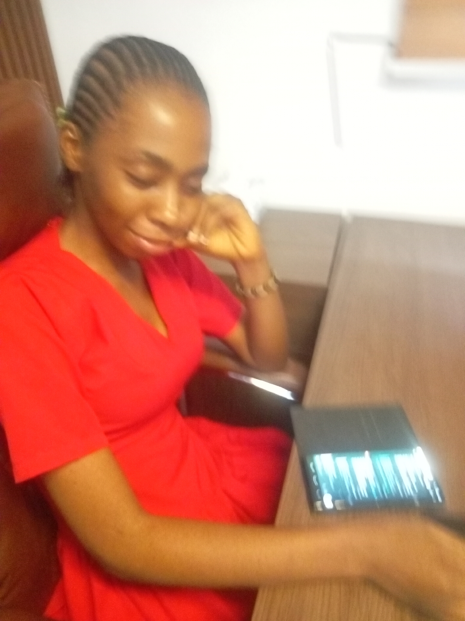 A woman with braided hair and wearing a red dress sits at a desk. Her head rests on her hand, and a tablet is on the desk in front of her. The location is Unknown, Unknown.