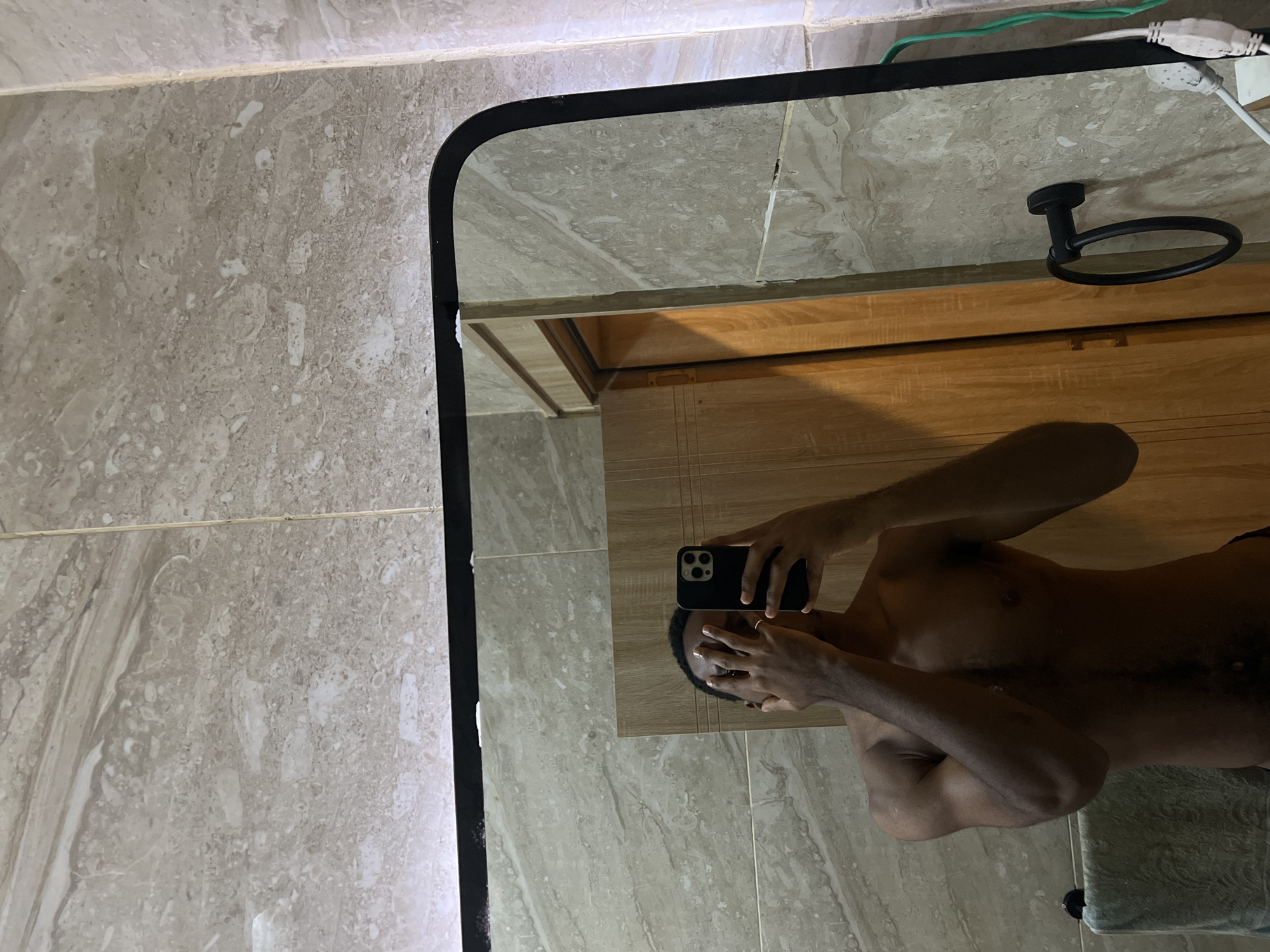 This image is a mirror selfie, rotated 90 degrees clockwise, depicting a person with dark skin. The person is reflected in a large mirror with a black frame, their upper torso and arms appearing horizontally across the lower part of the frame. They are shirtless and hold a black smartphone, likely an iPhone due to the visible camera array, to their face, obscuring their features with their hands and the device.
The setting appears to be a bathroom or dressing area, characterized by light brown or beige tiled surfaces with a mottled, marbled pattern. These tiles are visible directly on the wall to the left of the mirror and as reflections within the mirror itself. The reflection also shows a wooden cabinet or door positioned below the mirror, and a black, circular towel ring mounted on this wooden surface in the upper right portion of the mirror's view. In the top right corner of the mirror's reflection, green and white electrical wires are visible. A light-colored towel or fabric is partially visible at the bottom right of the reflection.
The lighting appears to be artificial, casting subtle shadows. There is no visible text in the image. The contextual location provided for this media is Gwarinpa, Nigeria.
