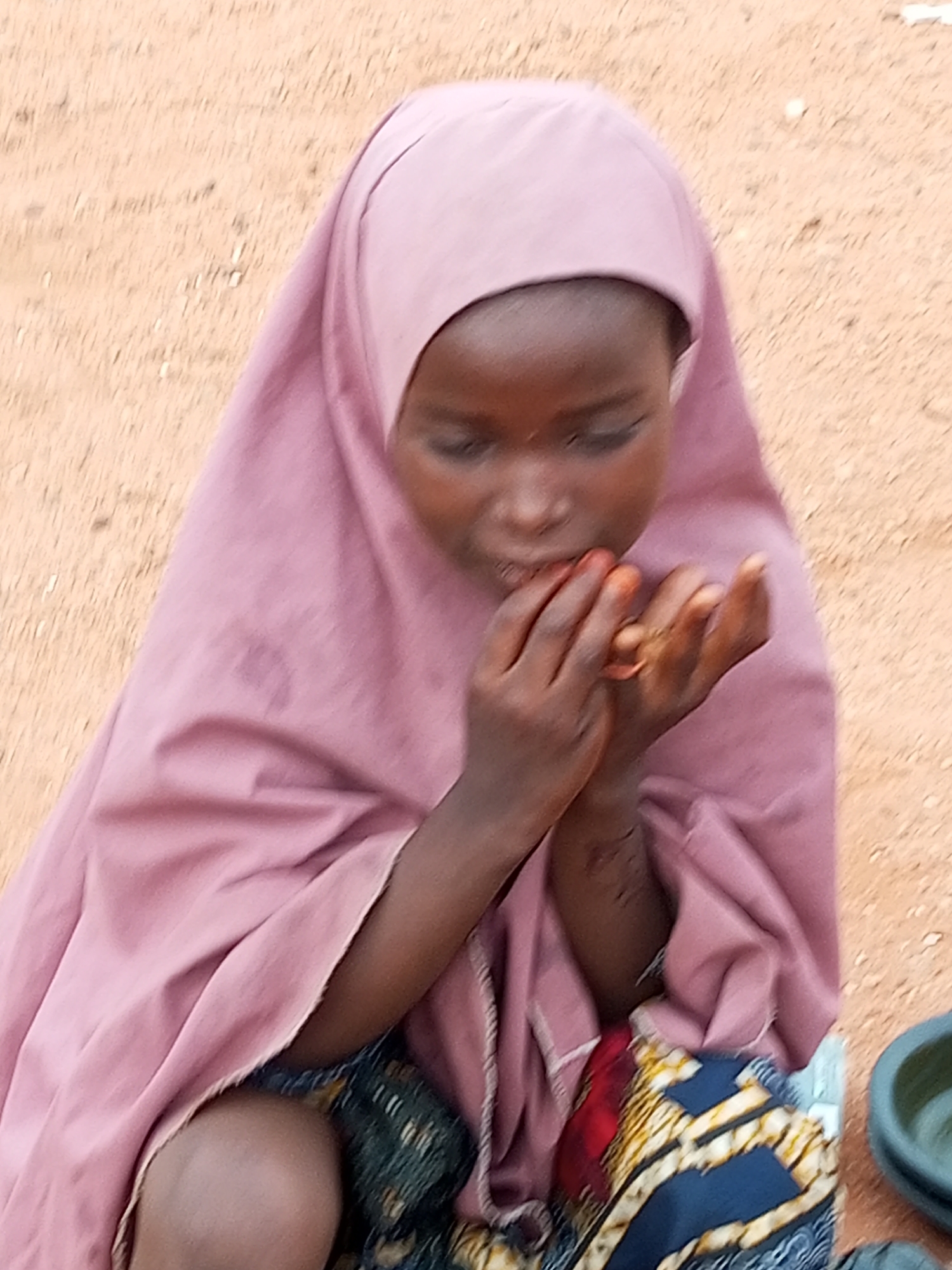In the city of Gombe, Nigeria, a young girl wearing a pink hijab and colorful fabric sits on the ground. Her hands are raised to her face, and she appears to be eating something small and reddish. The ground is sandy and appears to be dirt. The girl's expression is focused on her activity. The scene is likely outdoors, given the natural lighting and sandy environment.
