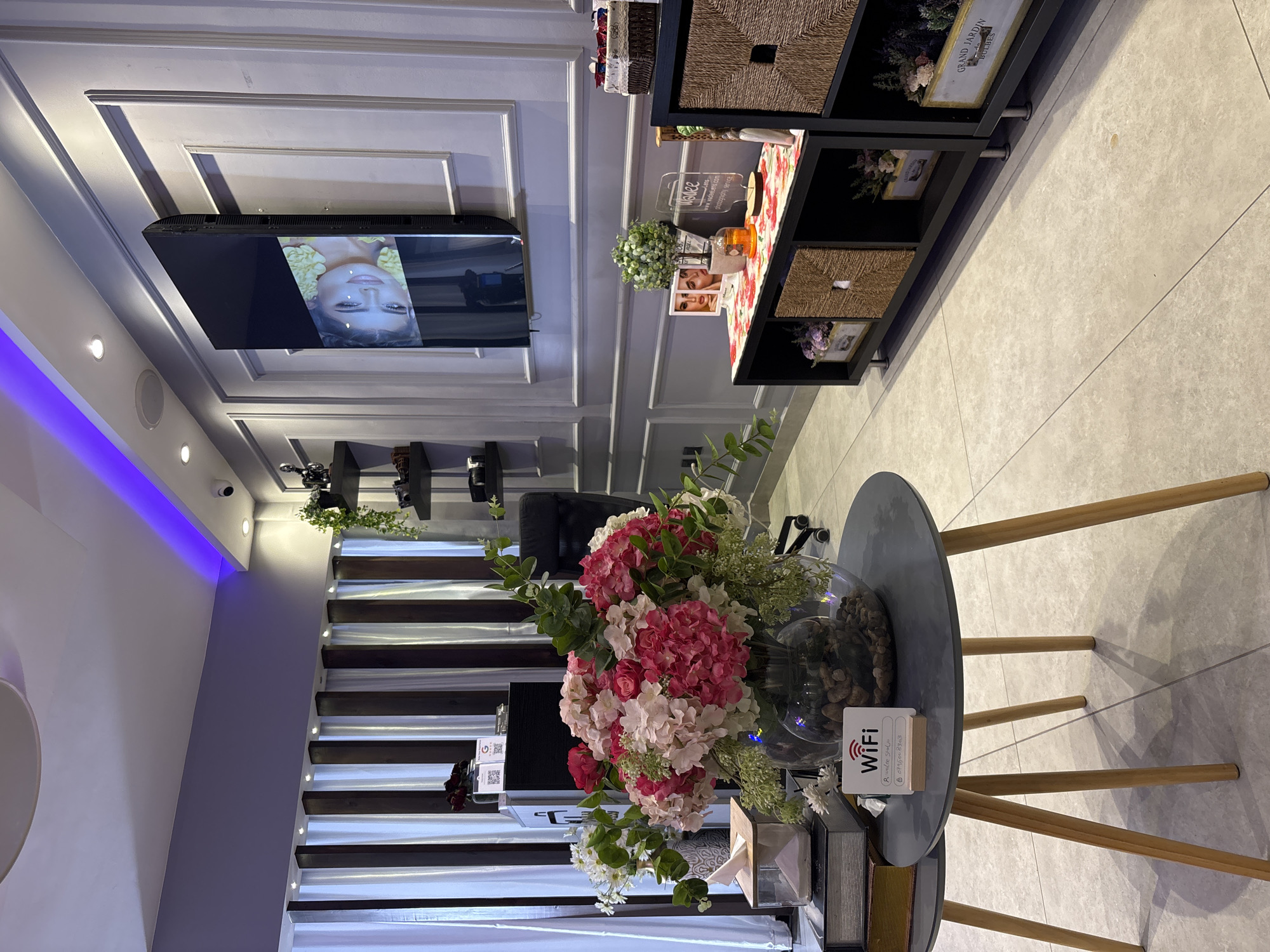 The image depicts an indoor scene, likely a reception area or waiting room in a salon or spa, given the aesthetic. The setting is modern and clean, with a gray tiled floor and white paneled walls. A large television screen is mounted on the wall, displaying a close-up of a woman's face with yellow flowers around her head. To the right of the television is a black shelving unit with woven baskets and decorative items, including small framed pictures and plants.
In the foreground, a round gray side table with wooden legs holds a large glass vase filled with a vibrant bouquet of pink and white hydrangeas and other greenery. Next to the vase, a small white sign with a QR code and the word "WiFi" is visible. There are also some decorative books and a tissue box on the table.
Behind the table, a staircase with dark wooden steps and illuminated edges leads upwards, suggesting a multi-level space. The lighting in the room is a mix of ambient and accent lighting, with a strip of purple light visible along the ceiling on the left side of the image. The overall mood is calm and inviting. There are no discernible cues about the time of day or weather, but the interior setting suggests it is not outdoors.
Visible text includes "WiFi" on the sign and text on a framed item on the shelf which appears to be a label for "GRAND JARDIN BULBES". There are also some smaller text elements on the QR code sign and on the television screen, but they are not legible.