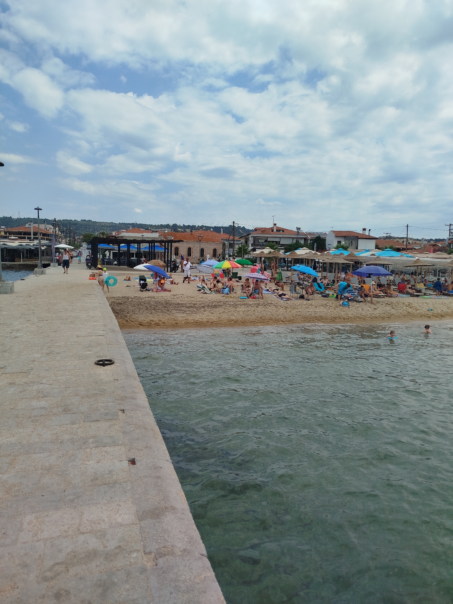 This image captures a bustling beach scene in Sithonia Municipal Unit, Greece. A stone promenade runs along the left side of the frame, leading towards a sandy beach packed with sunbathers. Beach umbrellas in various colors provide shade to the people lounging on towels and chairs. The water in the foreground appears calm and clear, with a few individuals swimming. In the background, a strip of buildings lines the coast, suggesting a well-developed tourist area. The sky is partly cloudy, with large white clouds drifting across a pale blue expanse. The overall atmosphere is one of leisure and activity, typical of a popular seaside destination.