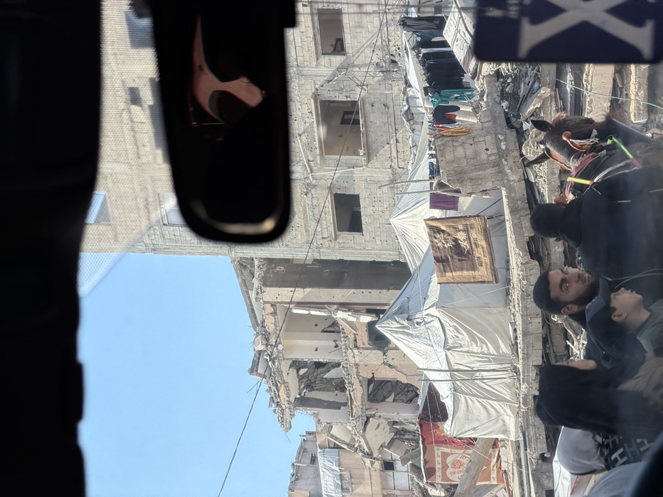 A damaged building, likely from conflict, dominates the view from inside a vehicle. The structure has visible holes and extensive debris. Makeshift coverings of white tarp are stretched across damaged areas. Clothes hang on lines strung between buildings. A group of people, including a man and a child, stand on the side. A dark object resembling a rearview mirror is visible in the foreground of the picture.