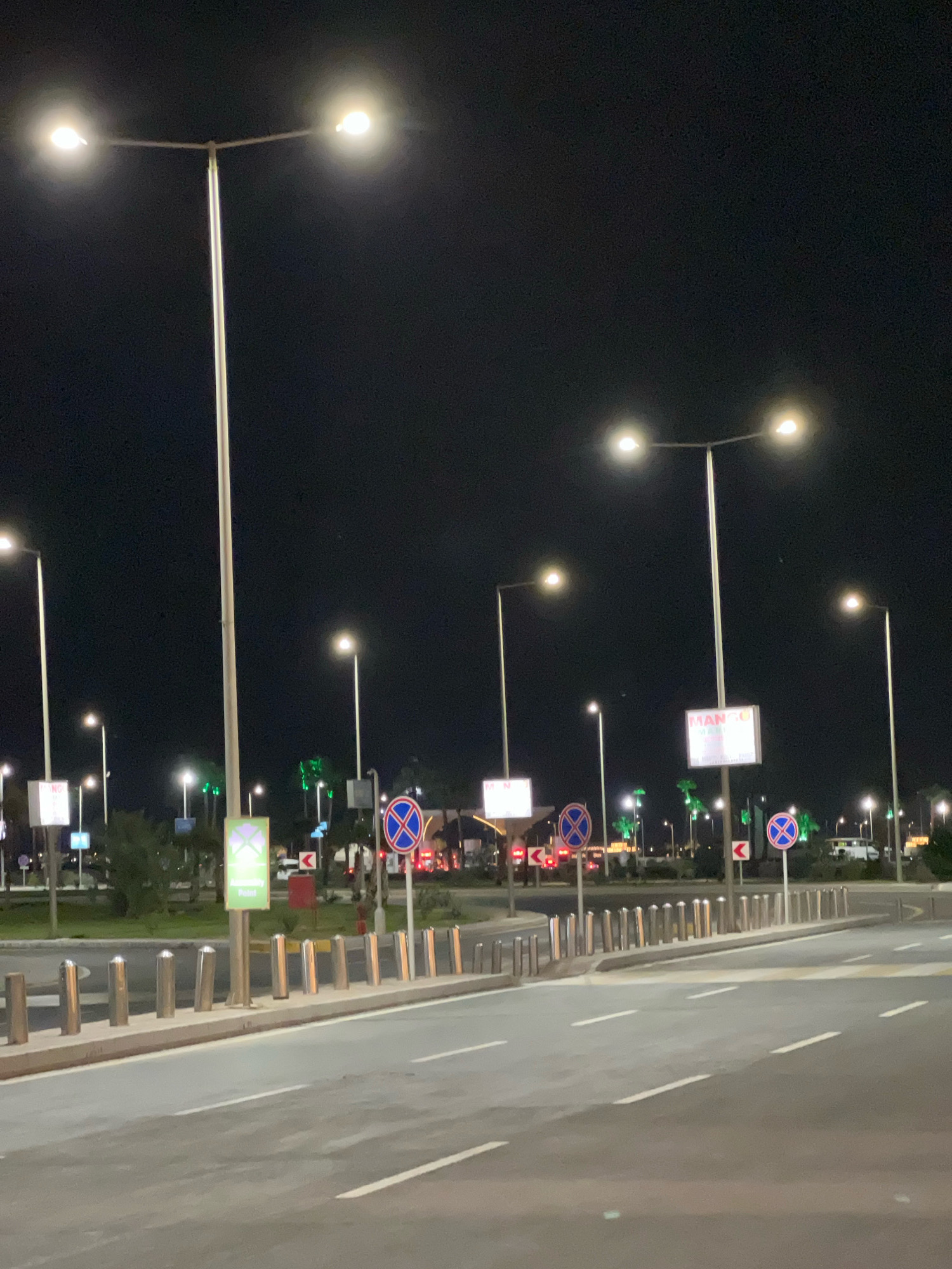 An outdoor nighttime scene in Sharm El Sheikh, Egypt, features a paved road with white lane markings, bordered by a row of polished metal bollards. Multiple streetlights with bright white bulbs illuminate the area. Several traffic signs are visible, including round blue signs with a red cross, indicating no parking or stopping, and a rectangular sign with a green symbol and text that appears to say "Accessibility Point." In the background, there are more streetlights and some trees, some of which are illuminated with green lights, possibly decorative. A few vehicles are visible in the distance. The sky is dark and clear.