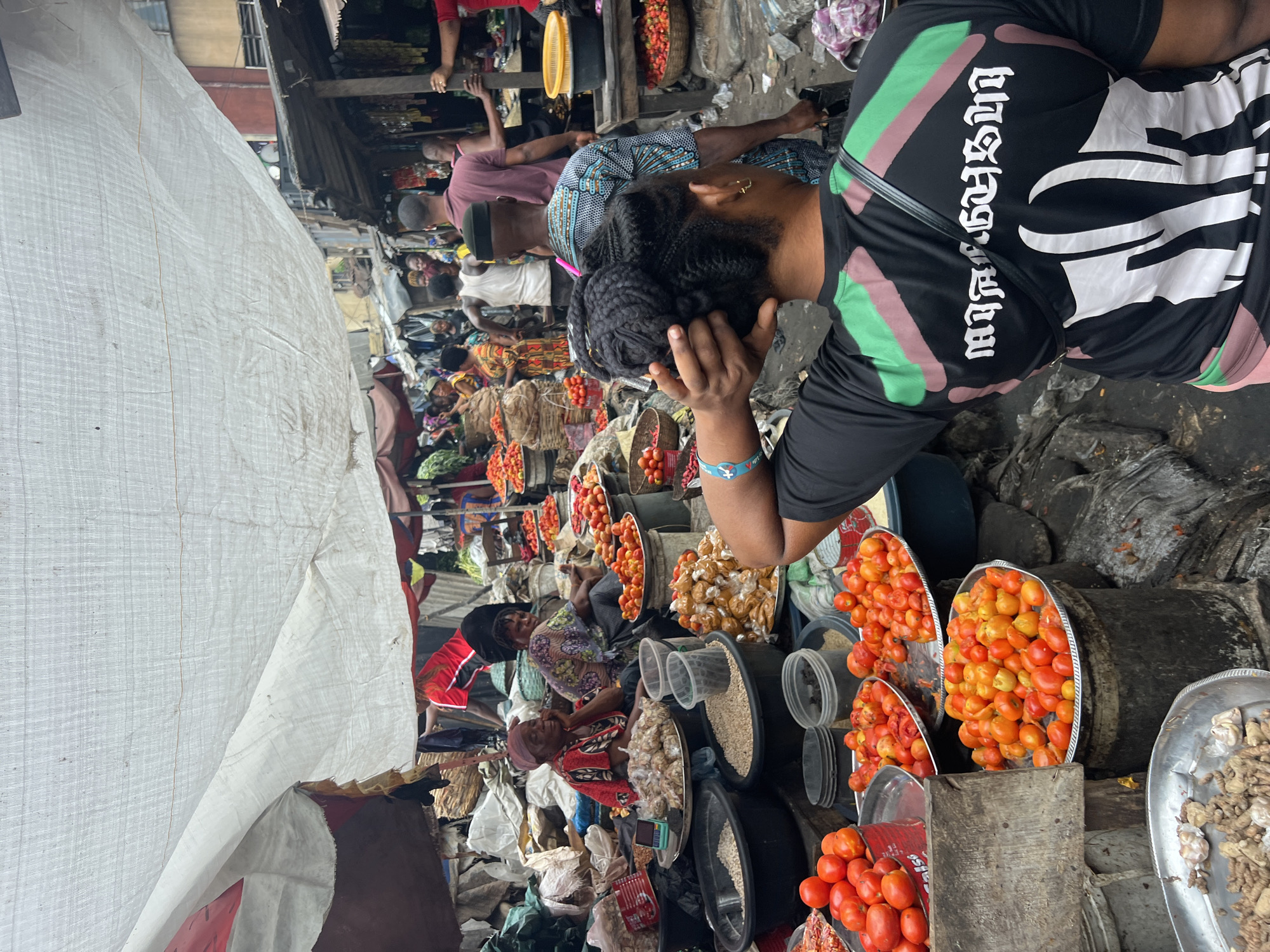The image depicts a bustling marketplace, likely in a West African city based on the appearance of the people and goods. The scene is dominated by vendors selling tomatoes, arranged in large metal trays and plastic containers. Other goods, possibly dried grains and spices, are visible in buckets and bags. Several people are present, including vendors and potential customers. The area is covered by a white tarpaulin, providing some shade. The overall environment appears crowded and potentially chaotic. The location is listed as Unknown, Unknown.