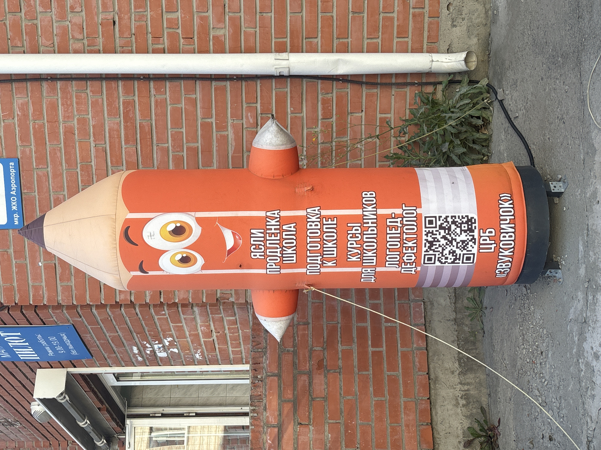 An inflatable orange pencil character with a smiling face stands on a paved surface in front of a brick building. The pencil has text on its body that reads "ЯСЛИ ПРОДЛЁНКА ШКОЛА ПОДГОТОВКА К ШКОЛЕ КУРСЫ ДЛЯ ШКОЛЬНИКОВ ЛОГОПЕД-ДЕФЕКТОЛОГ ЦРБ «ЗВУКОВИЧОК»" and a QR code. To the left of the pencil is a glass door and window of the building, with a blue sign above it that reads "ПИЛОТ" and "Режим работы: 9.00-19.00 (без выходных)". To the right of the pencil is a white pipe. The environment is outdoors, with some weeds growing near the base of the building. The building is constructed of red brick. The scene appears to be located in the city of Ob, Russia, as indicated by the blue sign.