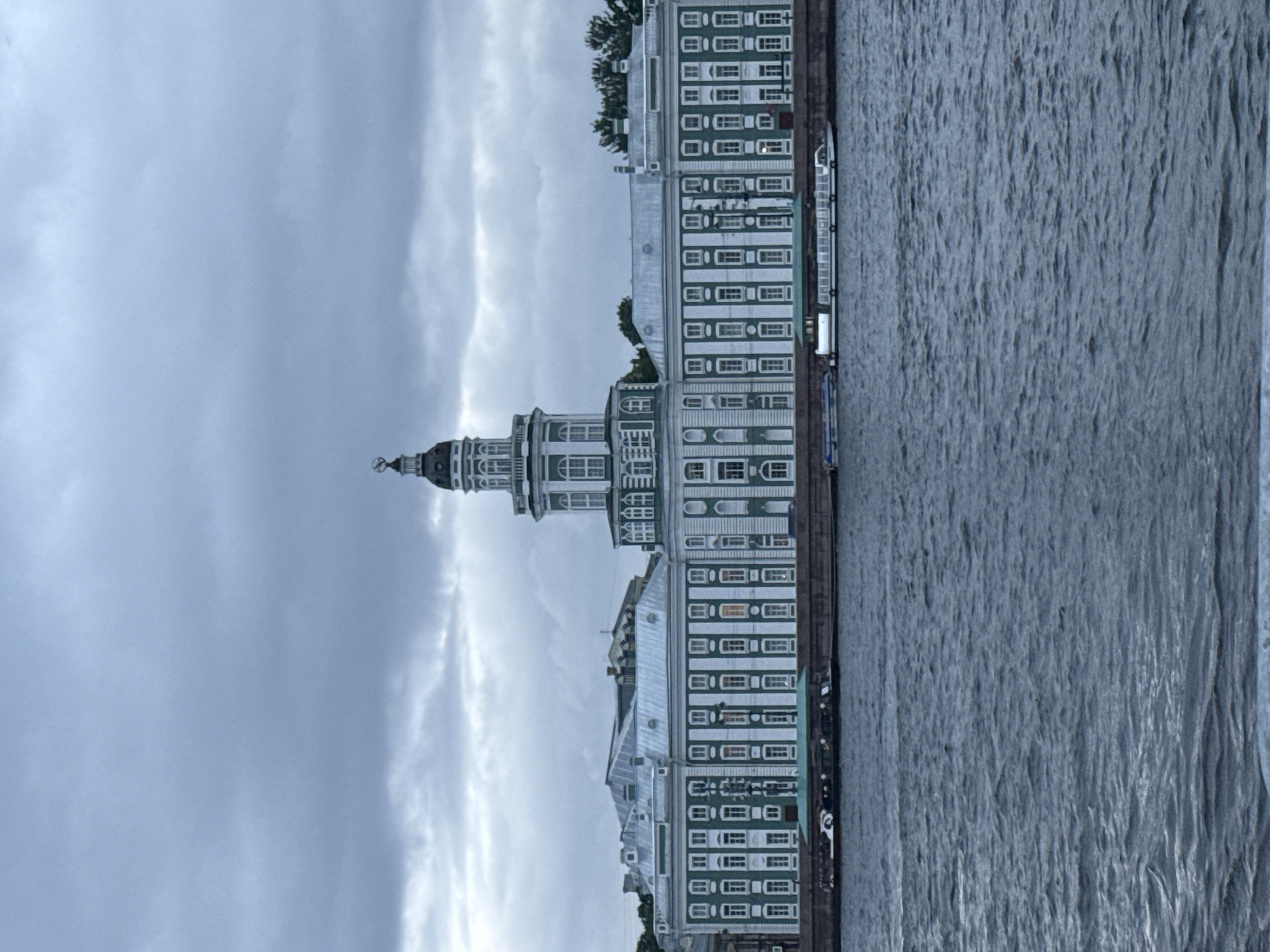 The image depicts a wide, choppy body of dark blue water in the foreground, likely a river or canal. In the background, across the water, is a large, historic building complex with a prominent central tower. The building has a light gray roof and a facade that is predominantly green and white, with numerous windows. The architecture suggests a classical or baroque style. Several boats are visible docked or navigating along the waterfront in front of the building. The sky above is overcast with heavy, dark gray clouds, indicating a gloomy or possibly stormy atmosphere. The overall scene is consistent with a view of the Kunstkamera museum, located on the Neva River in Saint Petersburg, Russia, known for its distinctive tower.