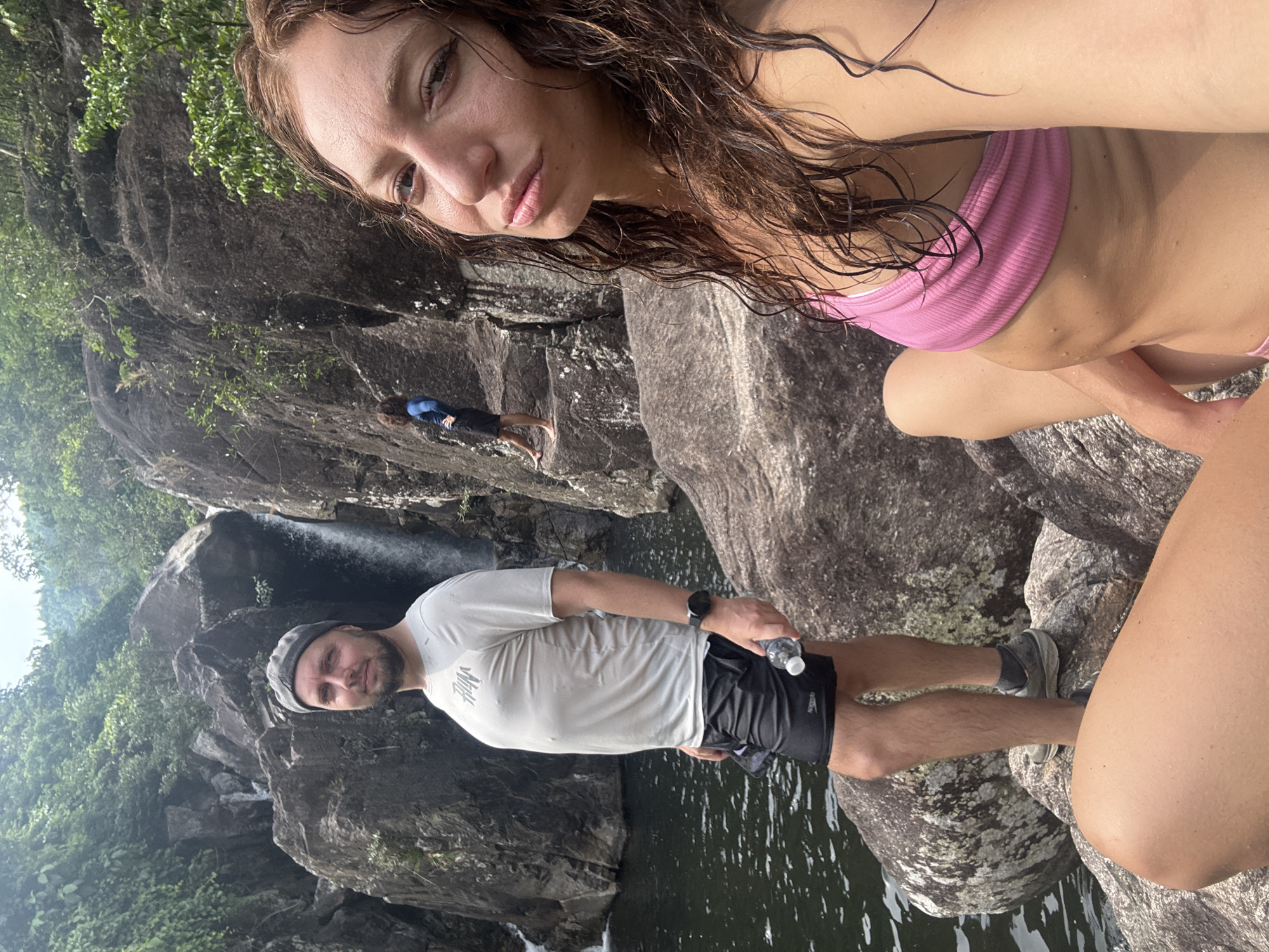 The image shows a selfie taken at a waterfall in Đà Nẵng, Vietnam. A woman with wet brown hair and a pink bikini top occupies the upper right of the frame. A man in a white t-shirt, black shorts, grey shoes, and a cap stands by the water's edge, holding a water bottle. Behind him is a waterfall flowing into a body of water surrounded by rocks and green vegetation. In the background, another person is seen climbing on the rocks.