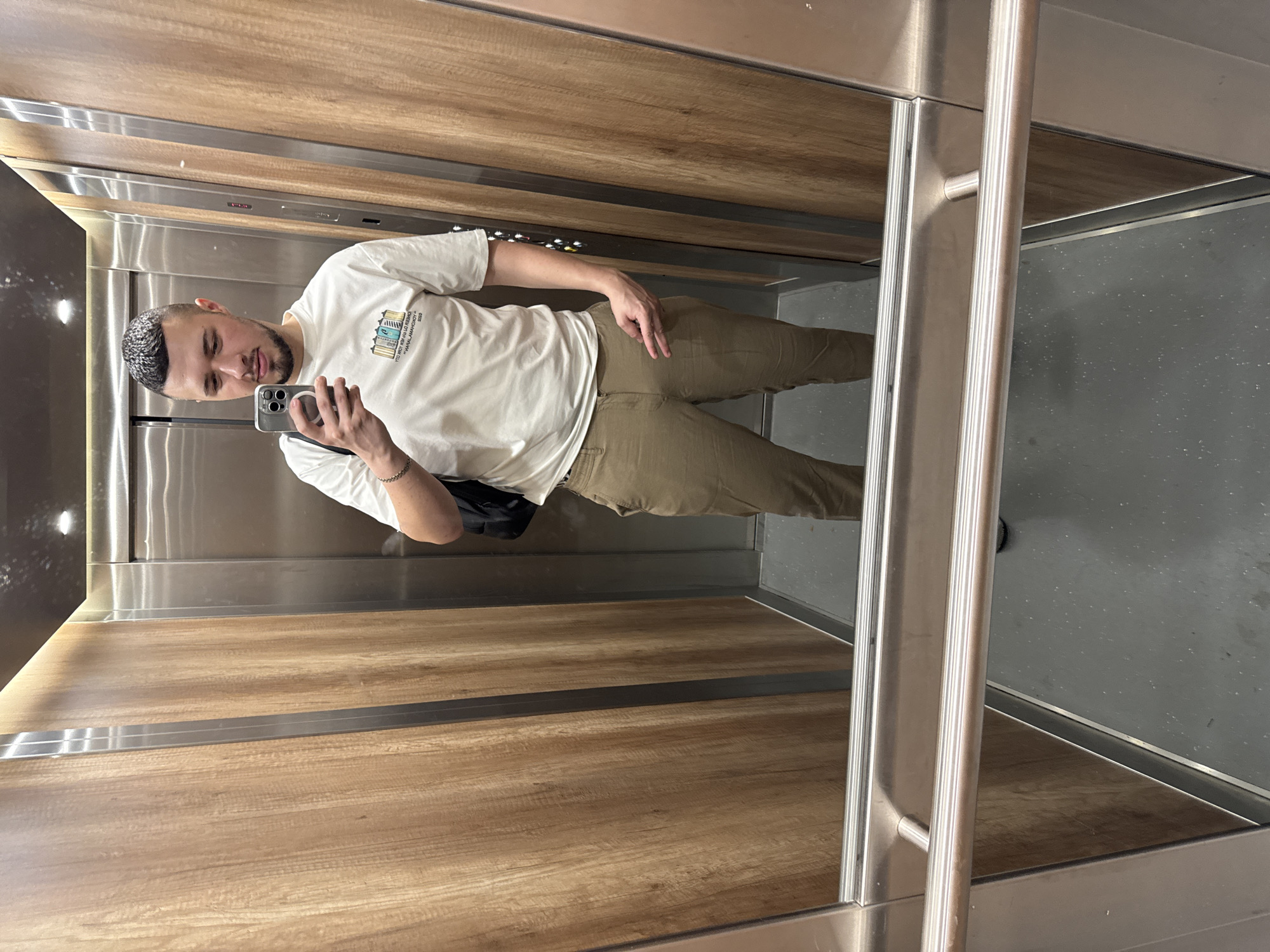 A man is taking a selfie in an elevator. The elevator's walls are wood-paneled on one side and grey on the other. The man is wearing a white t-shirt and khaki pants. He is holding a smartphone in his left hand. He has short dark hair and a beard. A black bag is slung over his shoulder. The elevator doors are metal. The location cannot be determined from the image.