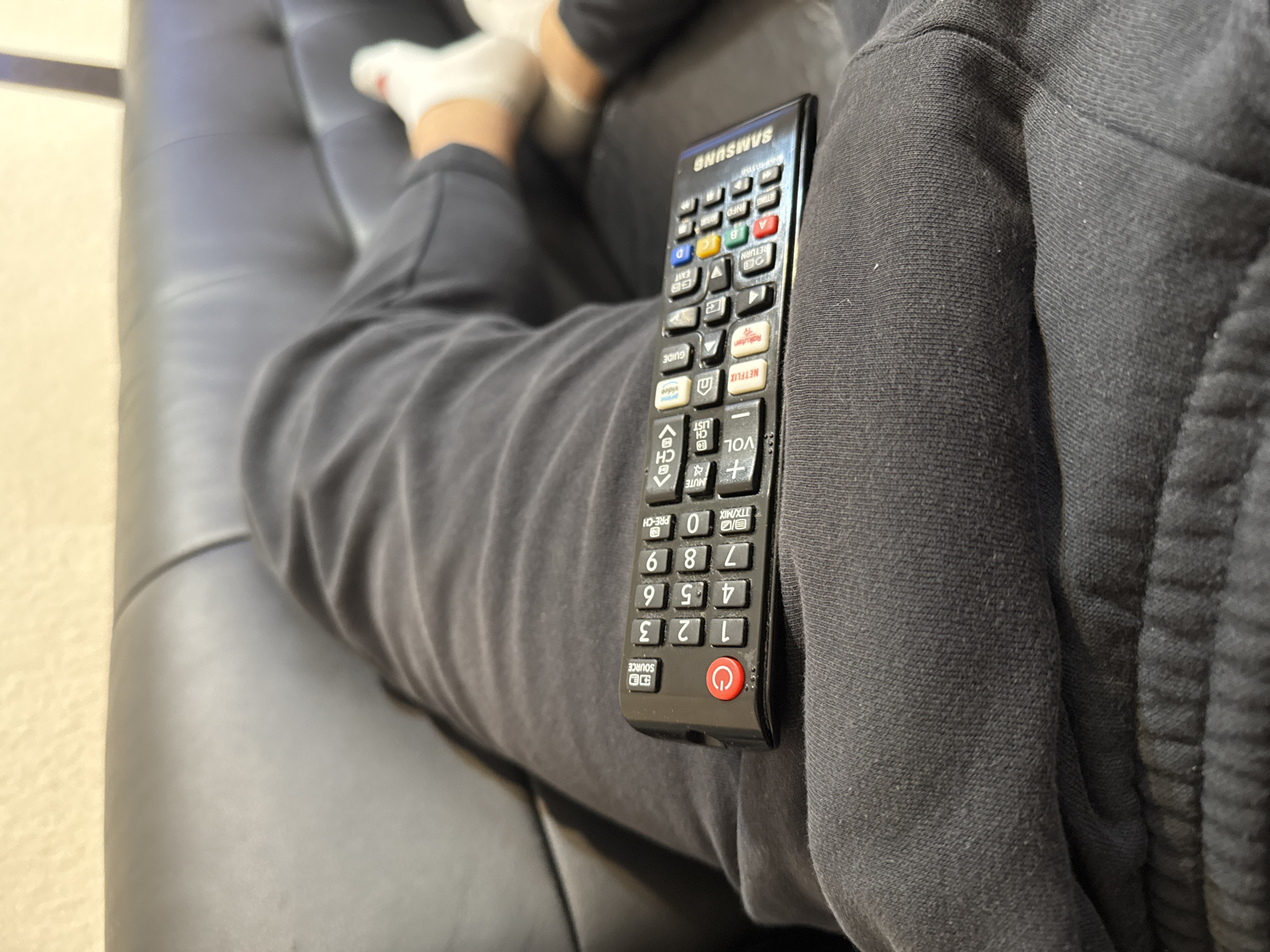 The image depicts a close-up, downward-angled view of a person reclining on a dark grey or black upholstered couch in an indoor setting, identified as the city of Delmenhorst, Germany. The individual is wearing dark grey sweatpants, which display visible fabric texture and seams, and white socks, partially visible on their feet in the background. A black Samsung television remote control is positioned on the person's upper thigh. The remote features a red power button, a numeric keypad (0-9), volume and channel controls, navigational buttons, and dedicated access buttons for streaming services such as Netflix and Rakuten TV. The background shows additional segments of the couch and a light-colored, textured wall.