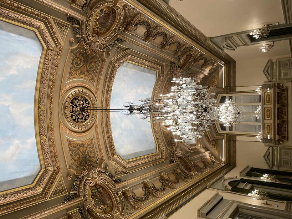 The image presents a lavish and highly ornate interior ceiling and the upper portions of its surrounding walls, indicative of grand European architecture in Paris, France.
The central feature is a magnificent, multi-tiered chandelier, crafted from sparkling clear glass or crystal, suspended from the ceiling and radiating a bright, diffused light. The ceiling itself is a complex arrangement of recessed panels, adorned with extensive gold leaf, intricate carvings, and elaborate decorative motifs including scrollwork, floral patterns, and acanthus leaves. Two large, central panels are painted with a serene blue sky and soft clouds, creating an illusion of open space. Integrated into the golden framework are circular medallions, some featuring sculpted faces or figures, and others containing small, detailed paintings or gilded patterns. A prominent, ornate circular grille in the ceiling structure serves as the mounting point for the main chandelier.
The upper sections of the walls continue the opulent design with decorative moldings and cornices. Large, tall windows or mirrors, framed with golden accents, reflect the abundant light, adding to the room's brilliance. Additional, smaller wall-mounted light fixtures (sconces) further illuminate the space.
The scene is static, devoid of people or any ongoing activity, allowing the viewer to fully appreciate the architectural and decorative artistry. The lavish use of gold, the classical motifs, and the scale of the decoration evoke a sense of historical grandeur and luxury, characteristic of a palace, opera house, or a prestigious building. No discernible text is visible in the image.