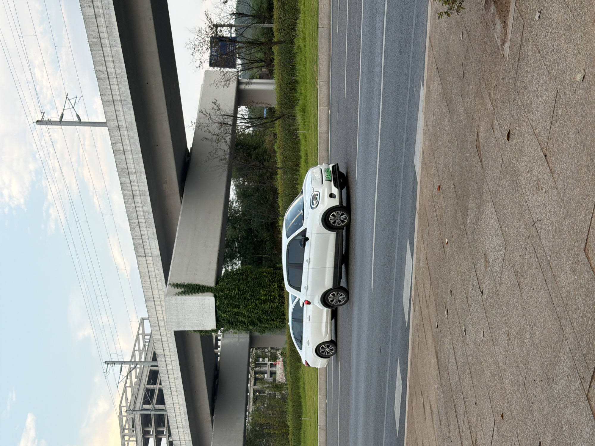 In Jiangjin, China, two white cars are parked side-by-side on an asphalt road. The car in the foreground is a compact SUV, facing right, with its license plate reading "A 331998". Behind it, a white sedan is parked, also facing right. Overhead, a concrete overpass supported by large pillars stretches across the scene. Green ivy climbs one of the pillars. The background features trees and a cloudy sky. A sidewalk made of stone tiles is visible in the lower portion of the image.