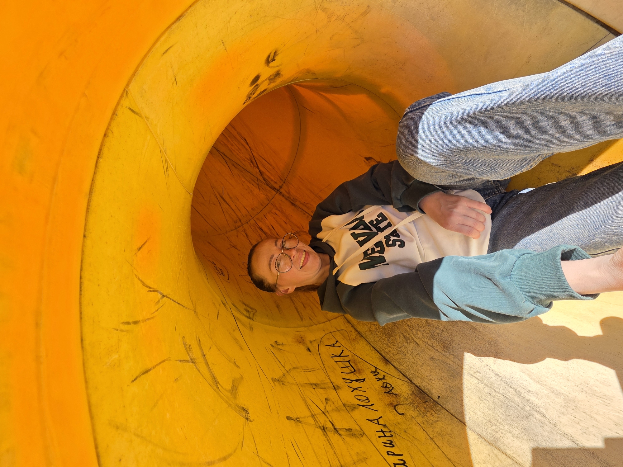 A young woman with round glasses is lying inside a large, bright orange playground tunnel. She is smiling and looking towards the camera. She wears a white hooded sweatshirt with black lettering, a turquoise green jacket, and light-wash denim jeans. The inside of the tunnel has graffiti in Russian writing. The tunnel appears to be located in Chaikovsky, Russia. The ground beneath the tunnel and beside the woman’s arm has a wooden surface with sunlight casting a shadow.
