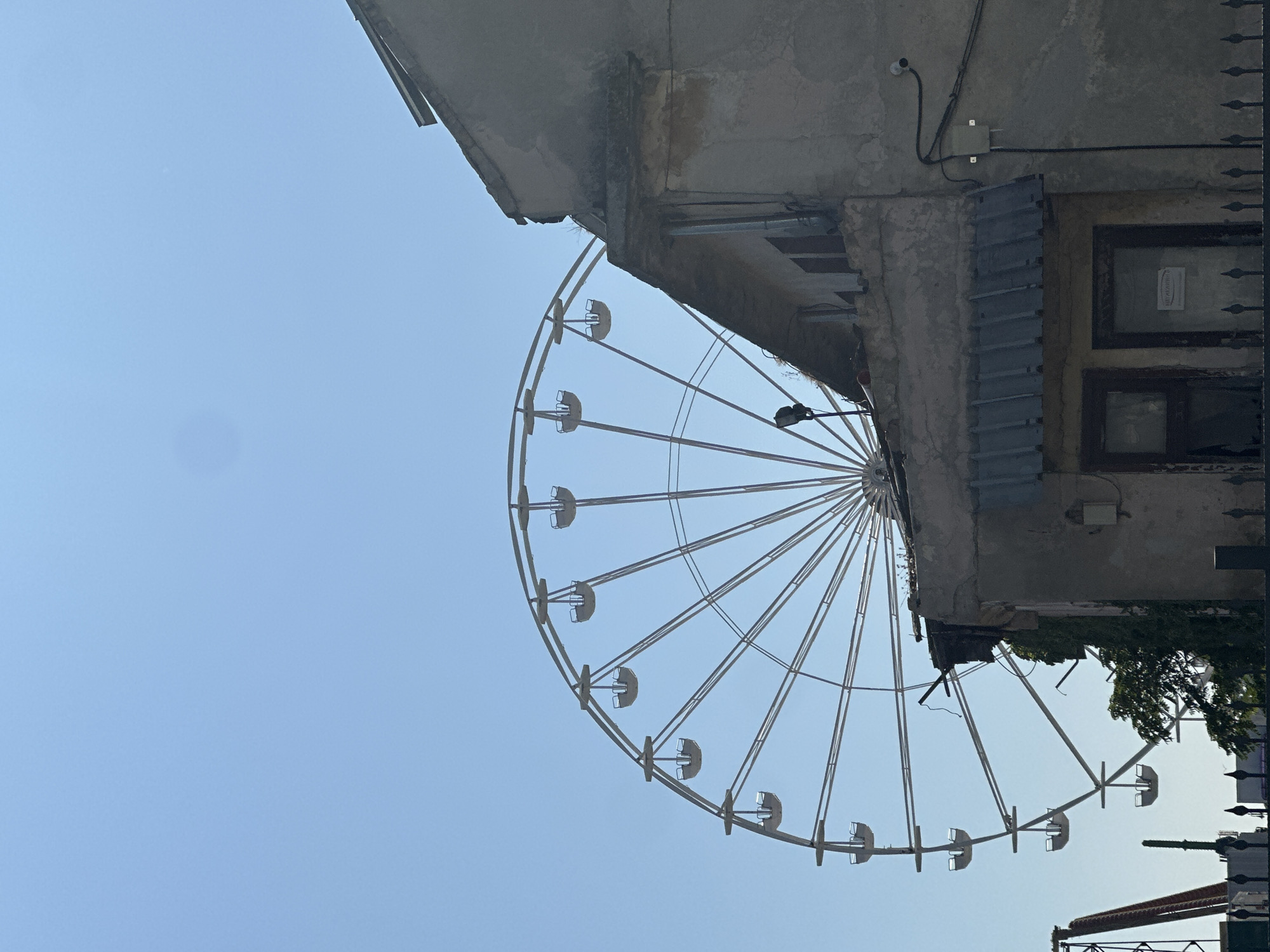 A Ferris wheel is visible in the background against a clear blue sky. In the foreground, an old, dilapidated building with a weathered facade and a damaged roof stands. The building has windows and what appears to be an awning. Electrical wires and a security camera are mounted on the exterior of the building. The scene is set in an urban environment, possibly an amusement park or a city area with older structures.