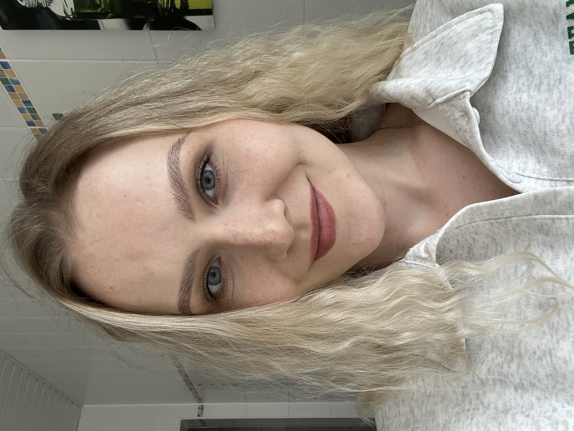 A close-up selfie features a young Caucasian woman with blonde hair and blue eyes. She is smiling and looking at the camera. The woman is wearing a light grey collared shirt. The background appears to be a bathroom with white tiled walls.