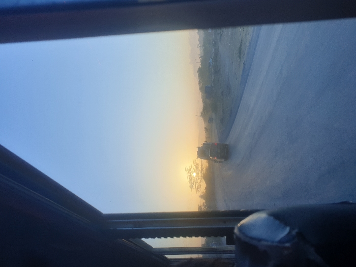 The image captures a view from inside a vehicle, looking out onto a road scene during either sunrise or sunset. The perspective is from a window or possibly a sunroof, indicated by the dark framing visible on the top, bottom, and partially on the left and right edges. The image is rotated counter-clockwise, making the horizon appear slanted.
The sky dominates the upper portion of the frame, transitioning from a soft, light blue in the top left to a brilliant golden-yellow and orange glow on the right, where the sun is low on the horizon. This strong backlighting silhouettes elements closer to the sun.
A paved road or highway stretches into the distance, occupying the mid-ground of the image. It appears somewhat dusty or hazy, especially as it recedes towards the horizon. On the left side of the road, a barren or sparsely vegetated landscape with indistinct distant structures or hills is visible. On the right side, closer to the bright sun, a cluster of trees or bushes stands out as dark silhouettes against the intense light.
Two vehicles, likely cars or SUVs, are visible on the road in the mid-distance, heading away from the viewer. They also appear as dark silhouettes against the bright backdrop, making their specific details indistinguishable. The time of day is clearly marked by the low sun and the warm, golden hues illuminating the scene, characteristic of either early morning (sunrise) or late afternoon/evening (sunset). The overall atmosphere has a hazy quality. Part of the vehicle's interior, possibly a hand or gloved finger resting on the window frame, is visible in the bottom right corner of the frame.