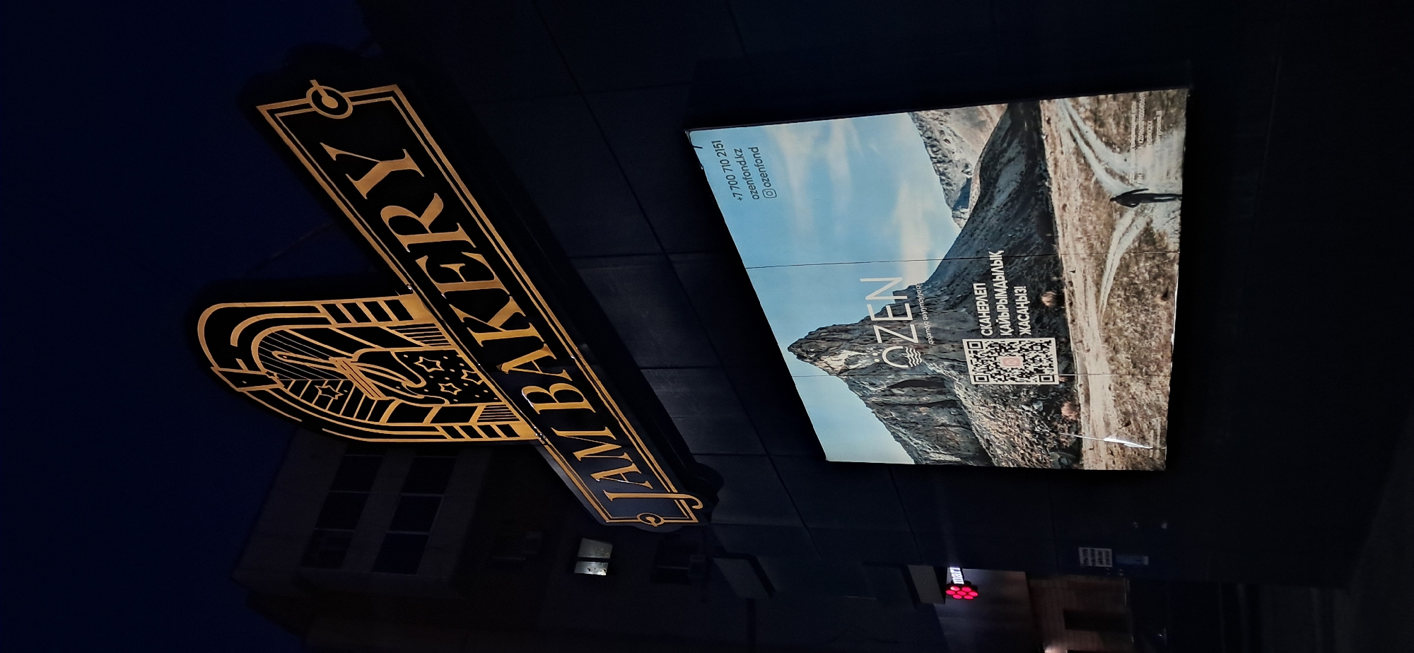 The image shows a street view at night, focusing on a large, illuminated sign for "JAM BAKERY" above a dark building facade. Below the bakery sign is a large digital billboard displaying a scenic landscape with a prominent mountain, a QR code, and some text in Kazakh. The billboard also includes contact information: a phone number "+7 700 710 2151", a website "ozenfond.kz", and an Instagram handle "@ozenfond". To the left of the billboard, a smaller red and white sign with the word "mart" is visible. The scene is dimly lit by artificial lights, with the sky appearing dark blue, suggesting it is nighttime. The overall impression is of an urban setting at night, likely a commercial area with businesses advertising their services.