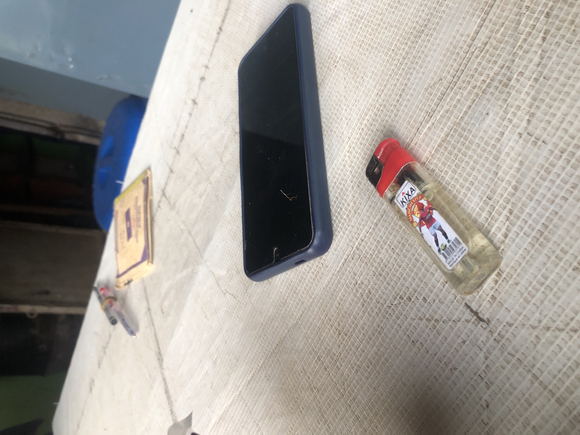 On a light-colored, textured surface, possibly a table, there are several objects. In the foreground, a transparent lighter with a red top and a Manchester United football player graphic is placed on its side. To the left of the lighter is a dark blue smartphone in a case, with its screen facing upwards and appearing black. Further up and to the left, a pen with a blue ink cartridge and a rubber band around its middle lies next to a small, folded yellow pamphlet or card. The background is blurred, showing indistinct dark shapes and a large blue object.