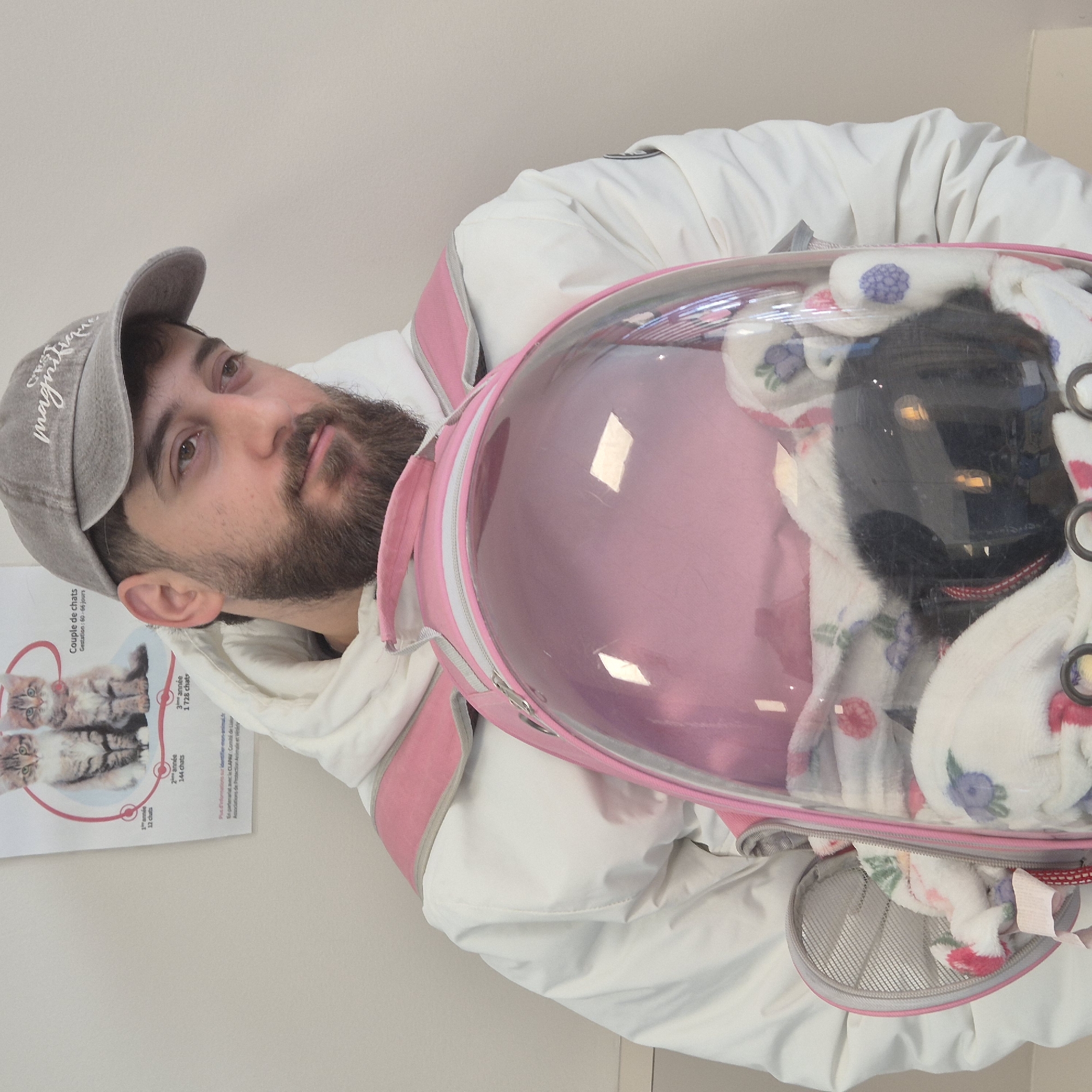 A man with a beard and a gray baseball cap holding a cat in a pink bubble backpack at a veterinary clinic in Geispolsheim, France. The man is wearing a white jacket. The cat is black and wrapped in a white blanket with floral patterns. There is a poster on the wall with information about cats.