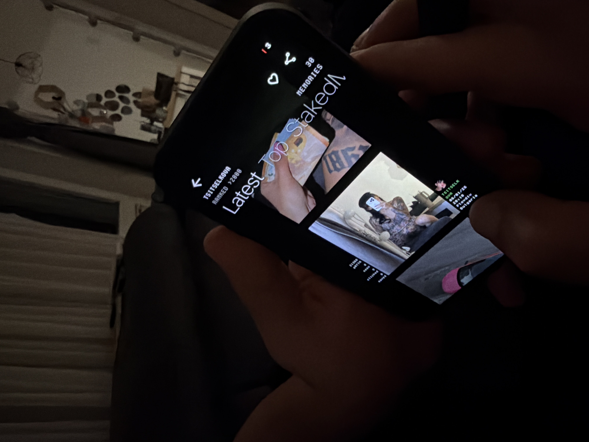 A close-up, low-light photograph shows a person holding a black smartphone with both hands, angled slightly to the left. The dimly lit background suggests an indoor setting, possibly a home, with a wall featuring decorative elements like small stones or artworks, and a horizontal fixture.
The smartphone screen is illuminated, displaying an application that resembles a social media or content-sharing platform. The prominent white text "Latest Top Staked" serves as a main title. Other visible text elements include "TSITSELKOVA," "RANKED >2008," and "MEMORIES." Icons such as a back arrow, a heart (like), and a share symbol are also present, accompanied by numbers "13" and "38," possibly indicating notifications or content counts.
The screen displays several thumbnail images. One shows a hand holding various banknotes, another features a person taking a selfie in a room, and a third reveals part of a bright pink shoe. Text associated with these posts includes "TSITSELK," "QUA," a date "06/01/26," a time "20:18," and location details "Plovdiv, Bulgaria," confirming the given context. Other small text fragments like "21368," "ORTTH," and "clicked" are also discernible, likely related to engagement metrics or post specifics. The overall low light conditions indicate the activity is taking place during the evening or night. The person's face and emotions are not visible.