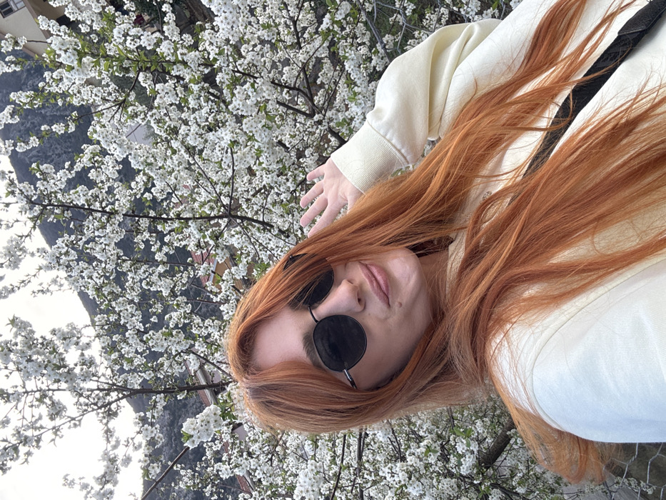 A female individual is captured in a close-up selfie against a backdrop of flowering trees and mountainous terrain in Škaljari, Montenegro. The subject, positioned centrally and in the foreground, has long, reddish-orange hair and wears dark, round-framed sunglasses. She is dressed in a light yellow or cream-colored long-sleeved sweatshirt, with a dark strap from a bag visible across her right shoulder. Her lips are slightly pursed in a subtle smile. Her left hand is raised, with fingers splayed and palm slightly visible.
The background features dense branches of a tree or shrub covered in numerous small white blossoms, indicative of spring. Behind the foliage, glimpses of residential buildings with light-colored facades and red-tiled roofs are visible on a hillside. Further back and higher in the frame, a segment of a rugged mountain or large hill is present, with varied darker and lighter tones suggesting natural rock and sparse vegetation. The sky is bright but mostly obscured by the flowering branches. The overall lighting suggests daylight conditions, possibly overcast.