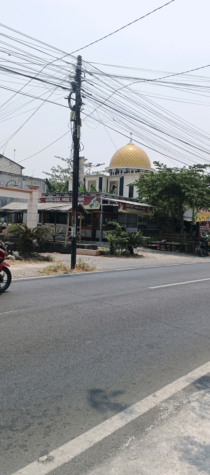 The image depicts a street scene in the city of Sumberwudi, Indonesia. A busy intersection is visible, with a prominent utility pole laden with numerous electrical and communication wires dominating the foreground. To the right, a golden-domed mosque stands out, its architecture featuring white walls and dark decorative elements. In front of the mosque, several small food stalls with outdoor seating are set up. A red motorcycle is partially visible on the left side of the road, and a portion of a parked motorcycle is seen on the far right. The road surface appears to be asphalt with white lane markings. The sky is overcast, suggesting a hazy or cloudy day. The overall environment appears to be a mix of urban infrastructure and commercial establishments.