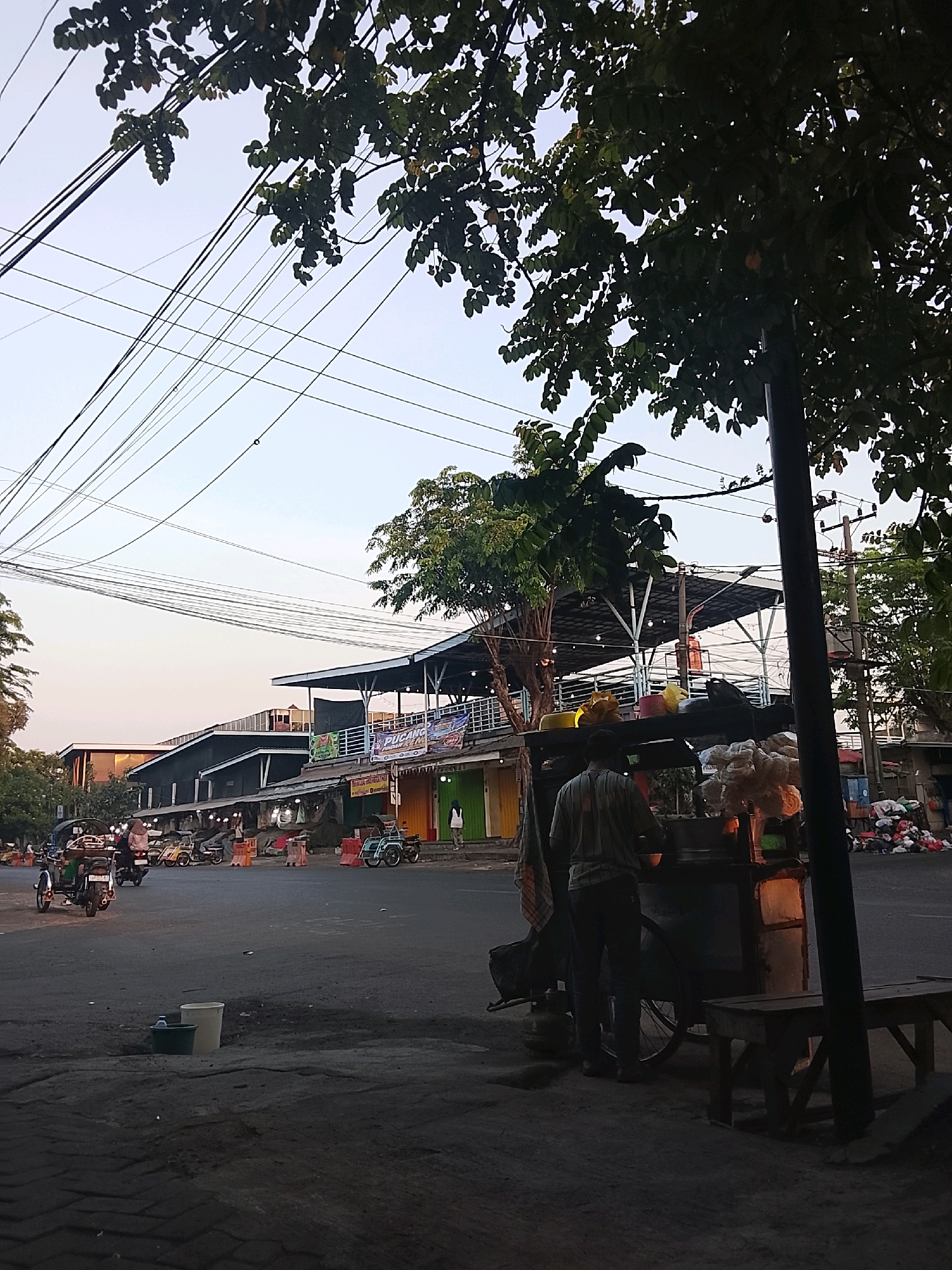 The image captures a street scene in Surabaya, Indonesia, likely in the late afternoon or early evening, judging by the lighting and shadows. The foreground shows a paved area with some debris and two buckets, one white and one green. A wooden bench sits on the right.
In the mid-ground, a food cart is set up on the side of a road. A person, wearing a striped shirt and dark pants, is standing behind the cart, appearing to be serving food. The cart itself is loaded with various items, including what looks like snacks or ingredients in plastic bags and containers. Another person is partially visible standing near the cart.
Behind the food cart, there is a row of buildings, possibly shops or market stalls, with awnings providing shade. Some of these buildings have signage, though the text is difficult to read. Several motorcycles and a small three-wheeled vehicle are parked or moving on the road.
The background features trees with dense foliage, and an overhead network of electrical wires crisscrosses the sky. The sky is a pale, hazy blue, suggesting a warm climate. The overall impression is a busy, everyday street scene in a bustling Indonesian city.