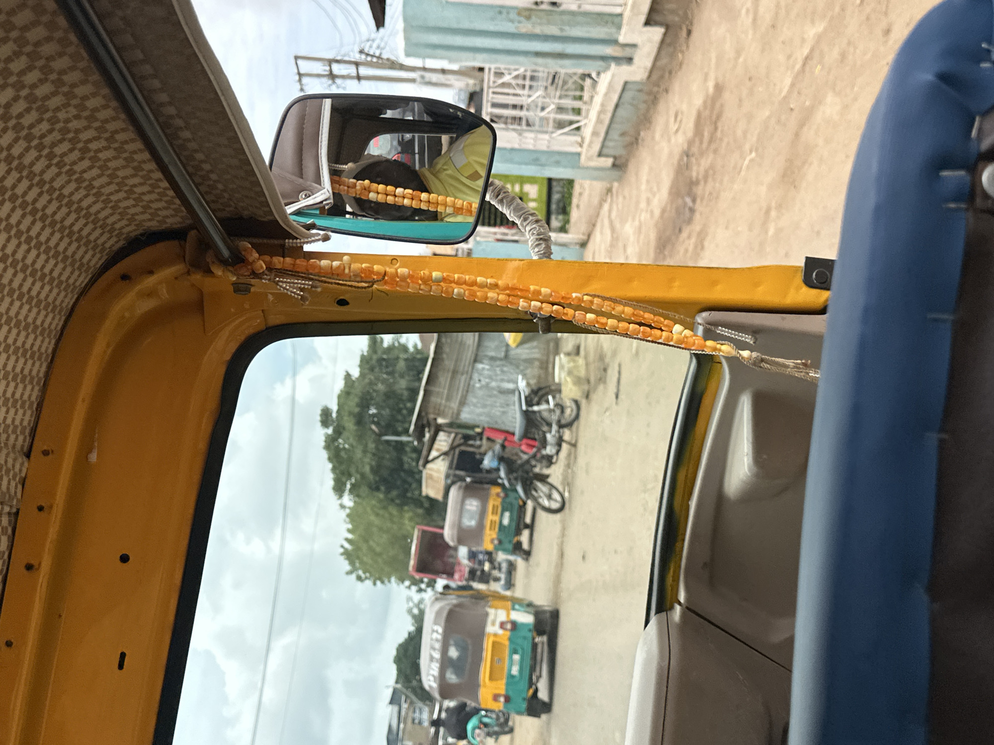 The image is taken from the interior of a yellow auto-rickshaw, looking outwards towards a dusty street in Jos, Nigeria. Through the windshield, several other auto-rickshaws and motorcycles are visible. The auto-rickshaw's interior features a checkered headliner and a blue cushioned seat in the foreground. A string of orange beads hangs from the interior structure. In the rearview mirror, the driver of the auto-rickshaw, wearing a yellow garment, is visible. The environment appears to be a busy street with low-rise buildings and trees in the background. The sky is overcast.