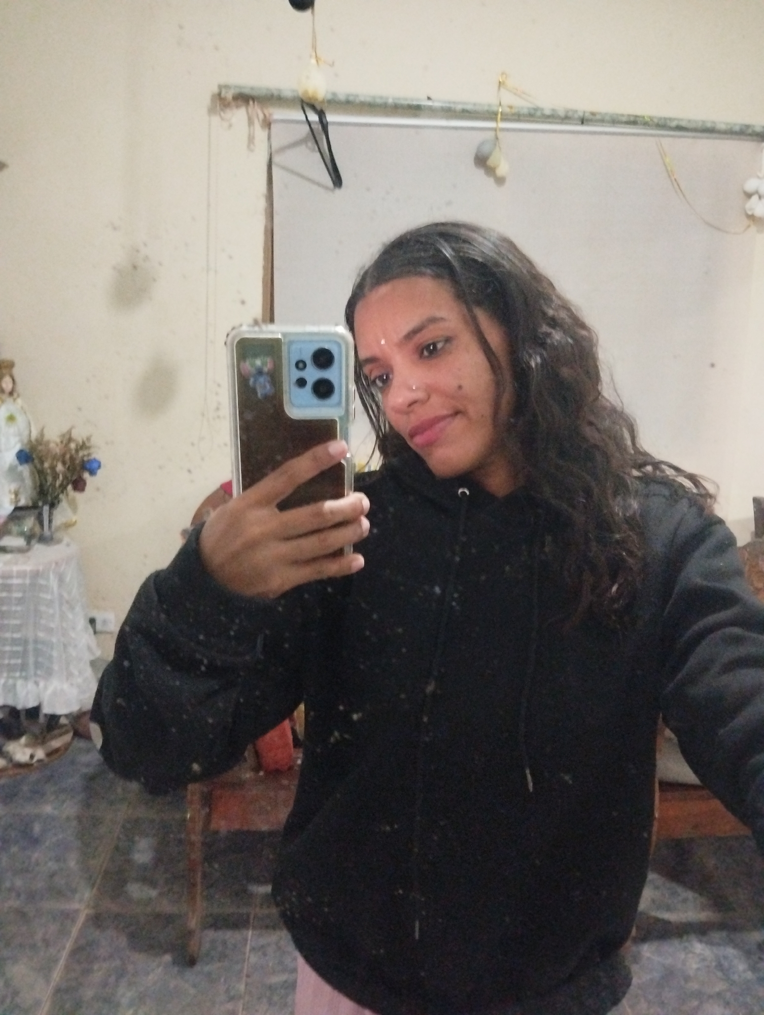 A woman is taking a selfie indoors in El Valle del Espíritu Santo, Venezuela. She is wearing a black hooded sweatshirt and has a nose ring and a mark on her forehead. She is holding a cell phone with a blue camera casing, and there is a reflection on the screen. In the background, there is a statue of a saint on a table with a white lace tablecloth, a mirror hanging on the wall, and a tiled floor. The wall is dirty.