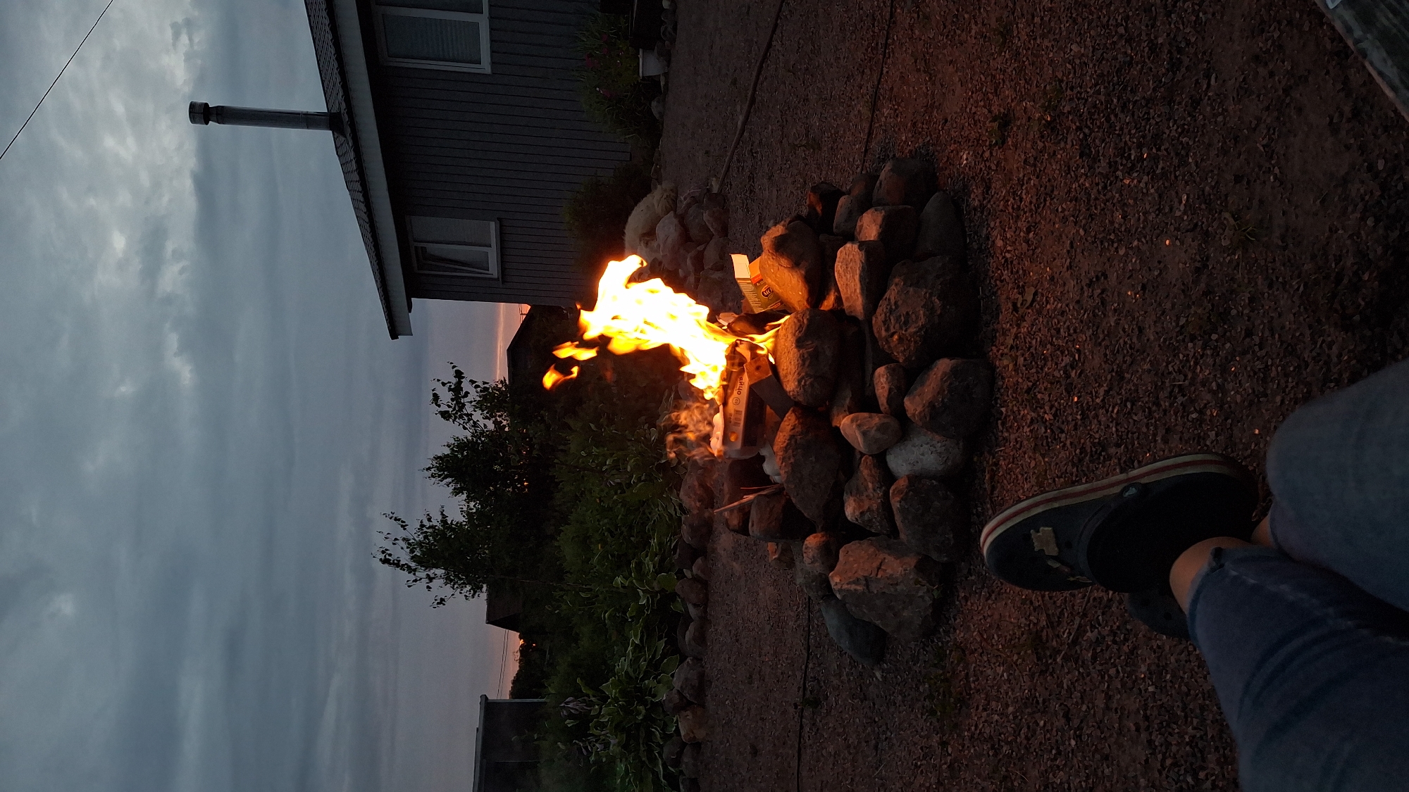 The image depicts a serene outdoor scene at dusk in Kempelevo, Russia. In the foreground, a person's legs and feet are visible, clad in blue jeans and black clogs, resting on a gravel-covered ground. A bonfire burns brightly in the center of the frame, surrounded by a ring of stones. The flames are vibrant orange and yellow, casting a warm glow on the rocks and the surrounding area. Behind the bonfire, a grey wooden house with white-trimmed windows and a chimney is partially visible, nestled among lush green vegetation. The sky above is overcast with a gradient of gray and hints of pink and orange from the setting sun. The overall atmosphere is peaceful and intimate, suggesting a moment of relaxation and connection with nature.