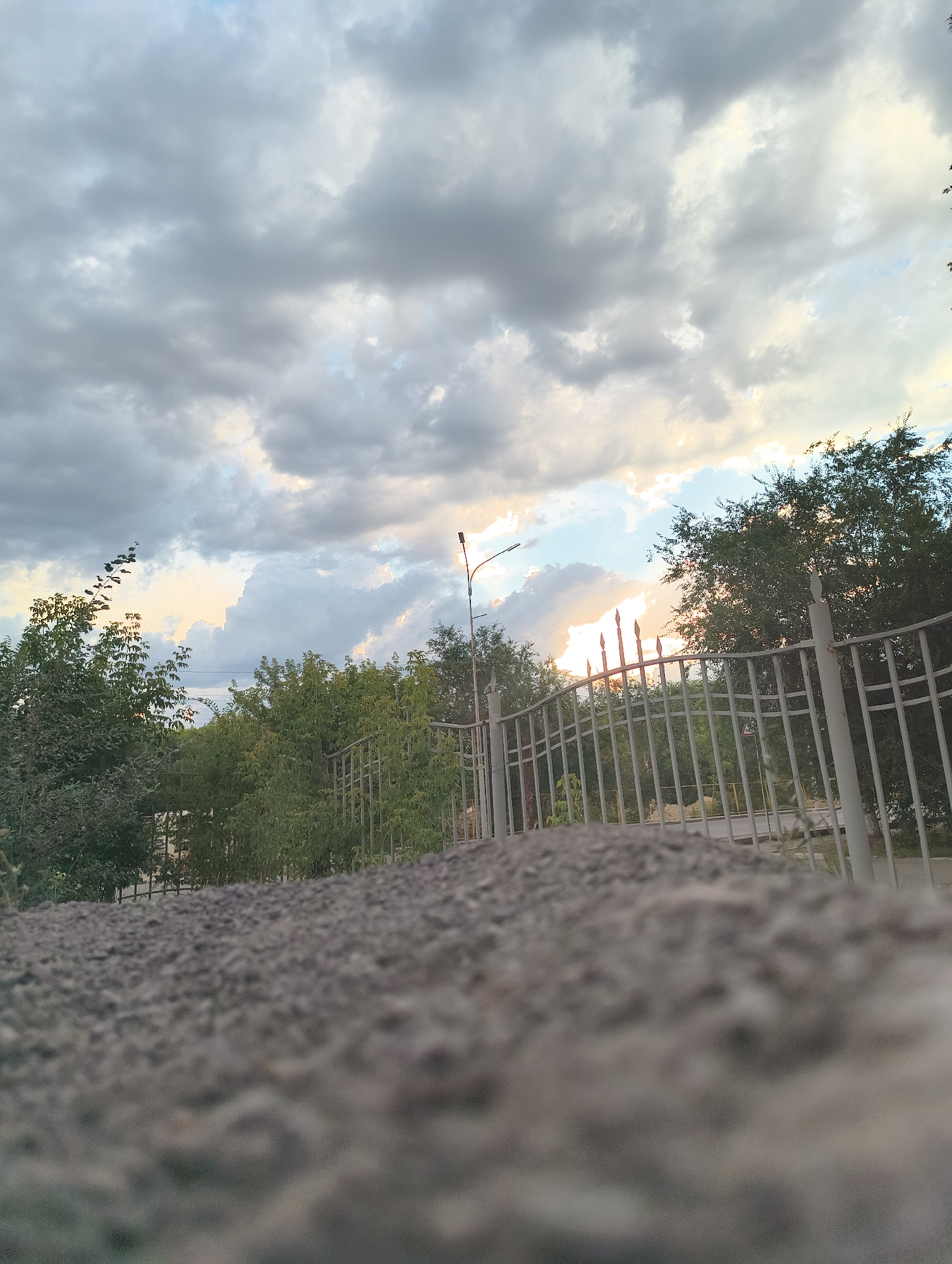 The image captures a view of a cloudy sky with sunlight breaking through the clouds. In the foreground, there is a textured surface of gravel and a light-colored metal fence. Beyond the fence, a line of trees is visible, with some of the trees appearing fuller and greener than others. A street lamp is also present on the right side of the image. The overall impression is of an outdoor scene, possibly in a park or residential area, during late afternoon or early evening.